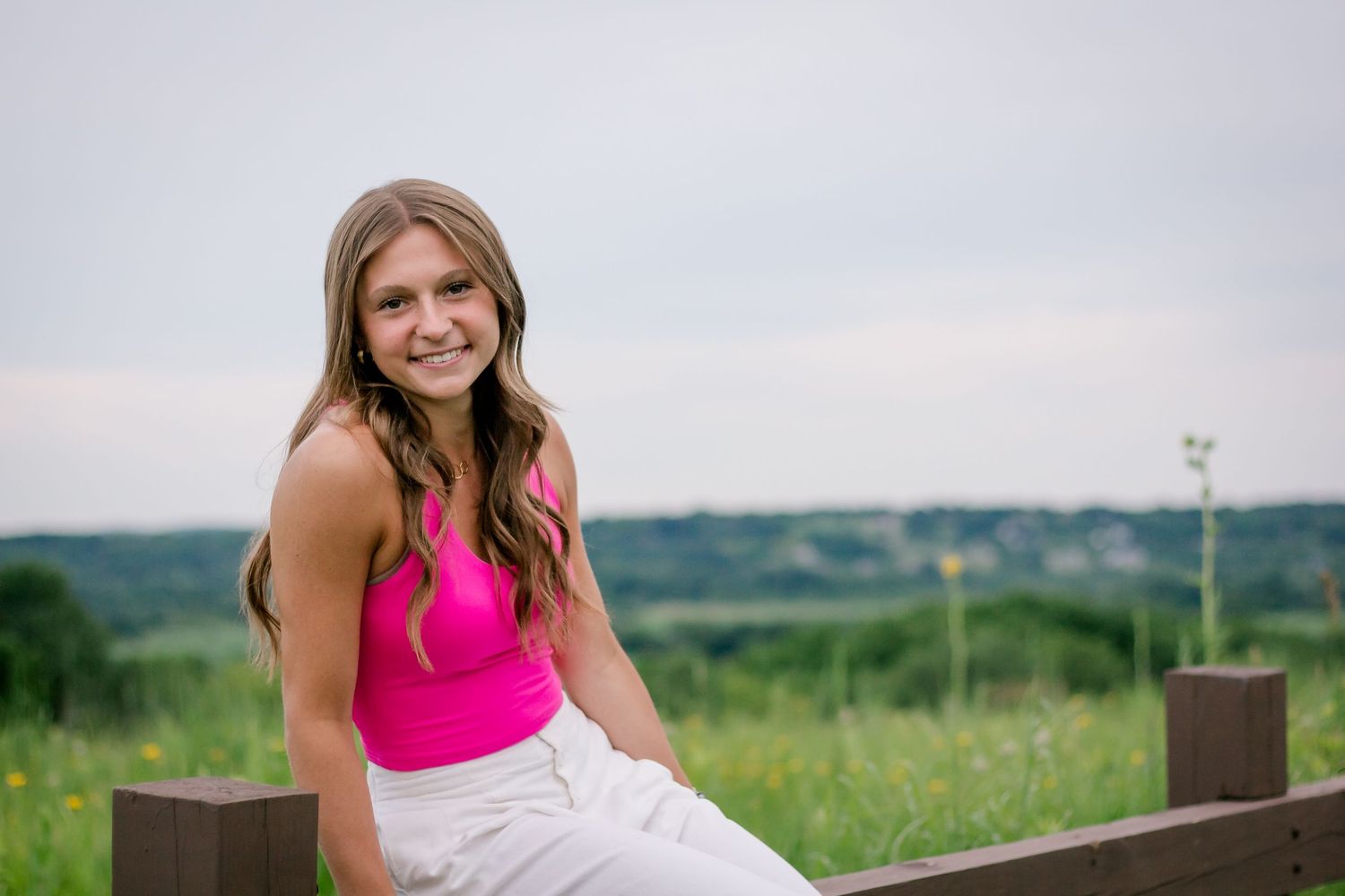 Athletic woman in pink tank top sits casually on wooden fence overlooking scenic countryside landscape.