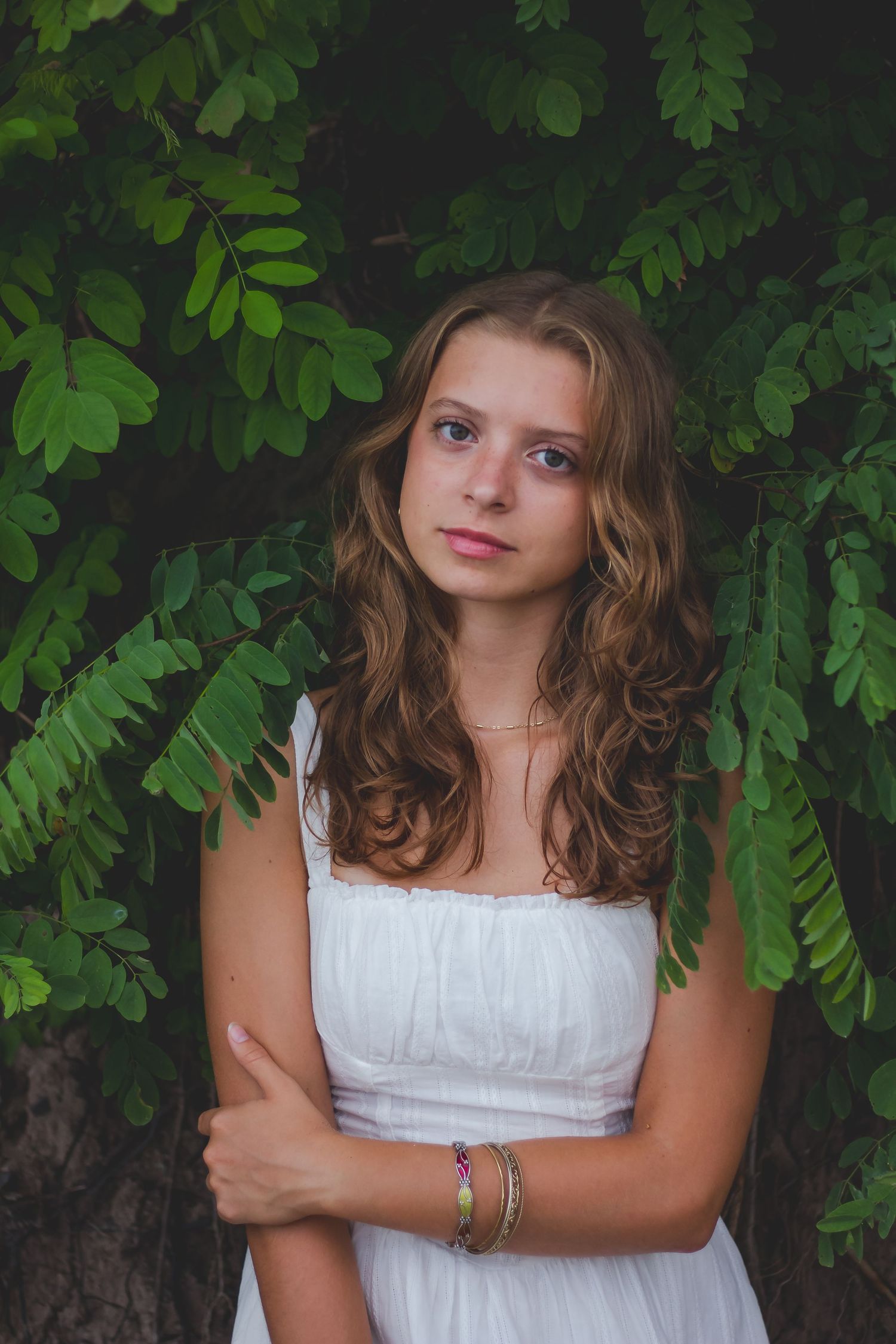 A person in a white dress poses against lush green foliage in a natural outdoor setting.