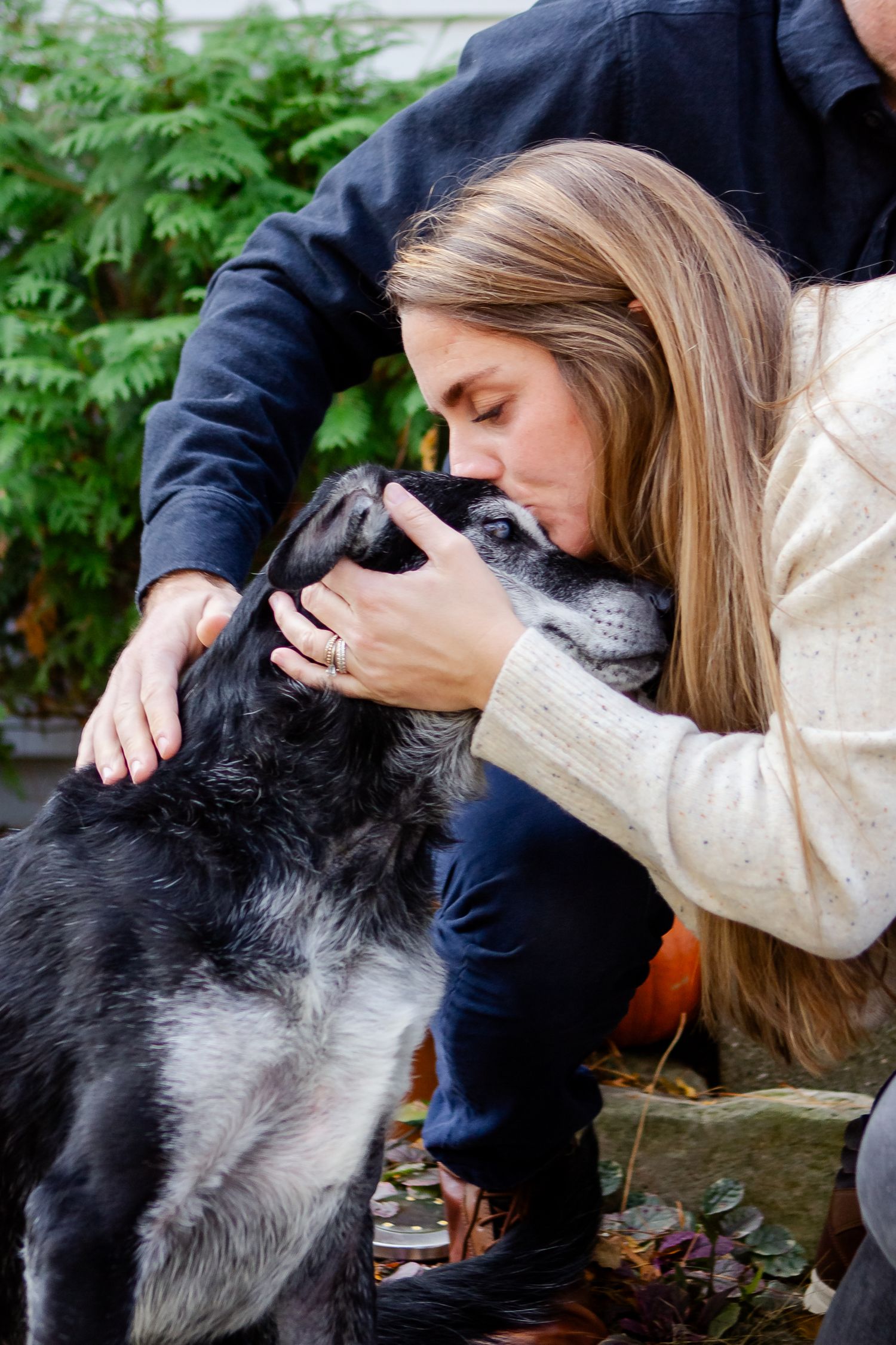 A person in a cream sweater lovingly pets and embraces a large black dog outdoors among green foliage.