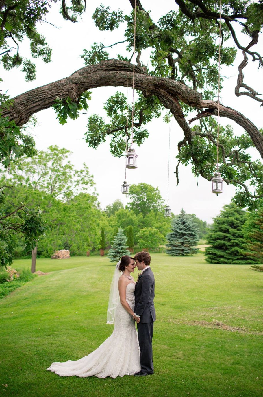 Wedding couple poses under a majestic oak tree with hanging lanterns in a garden setting.