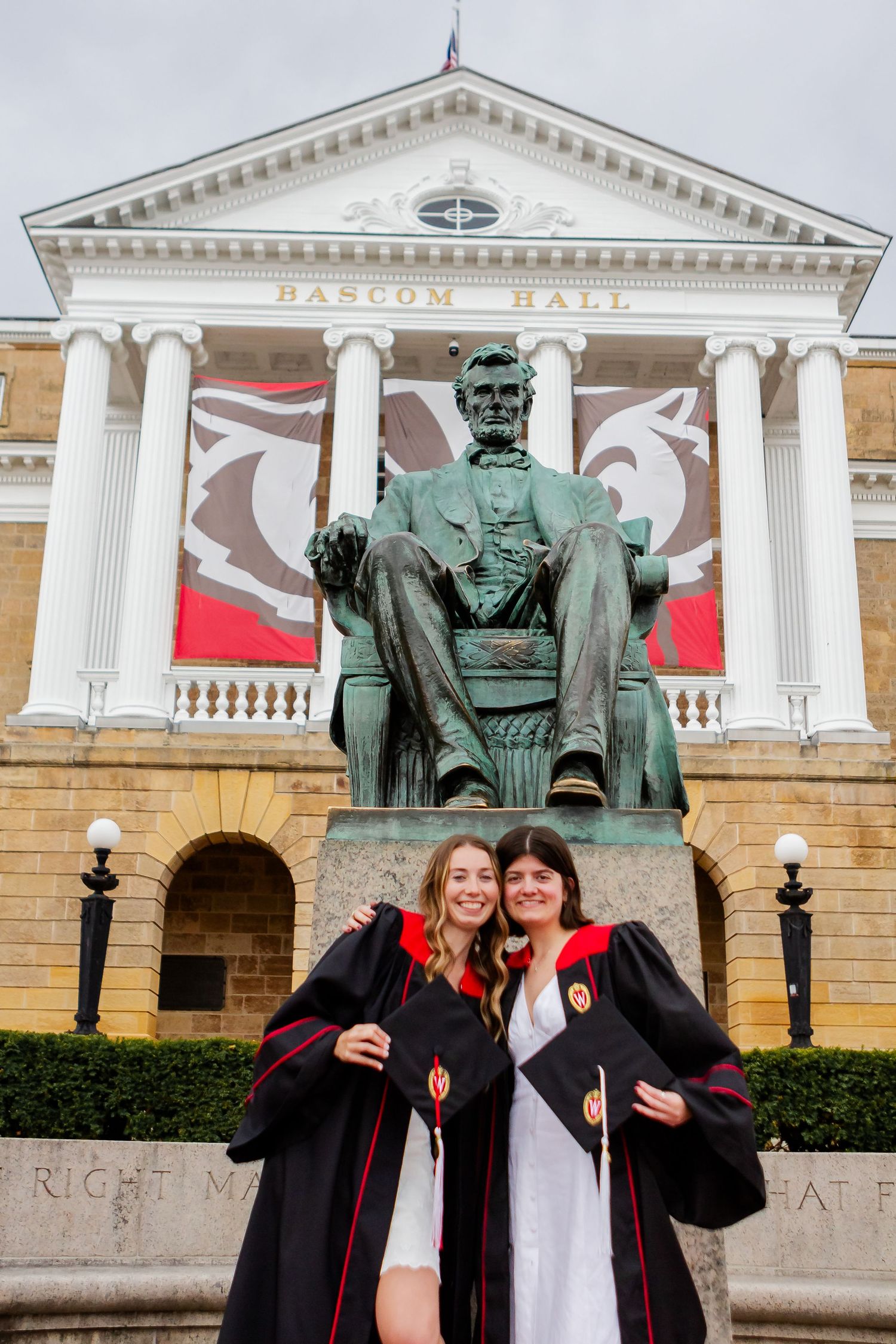 Graduates in black gowns celebrate in front of neoclassical university building with bronze statue.
