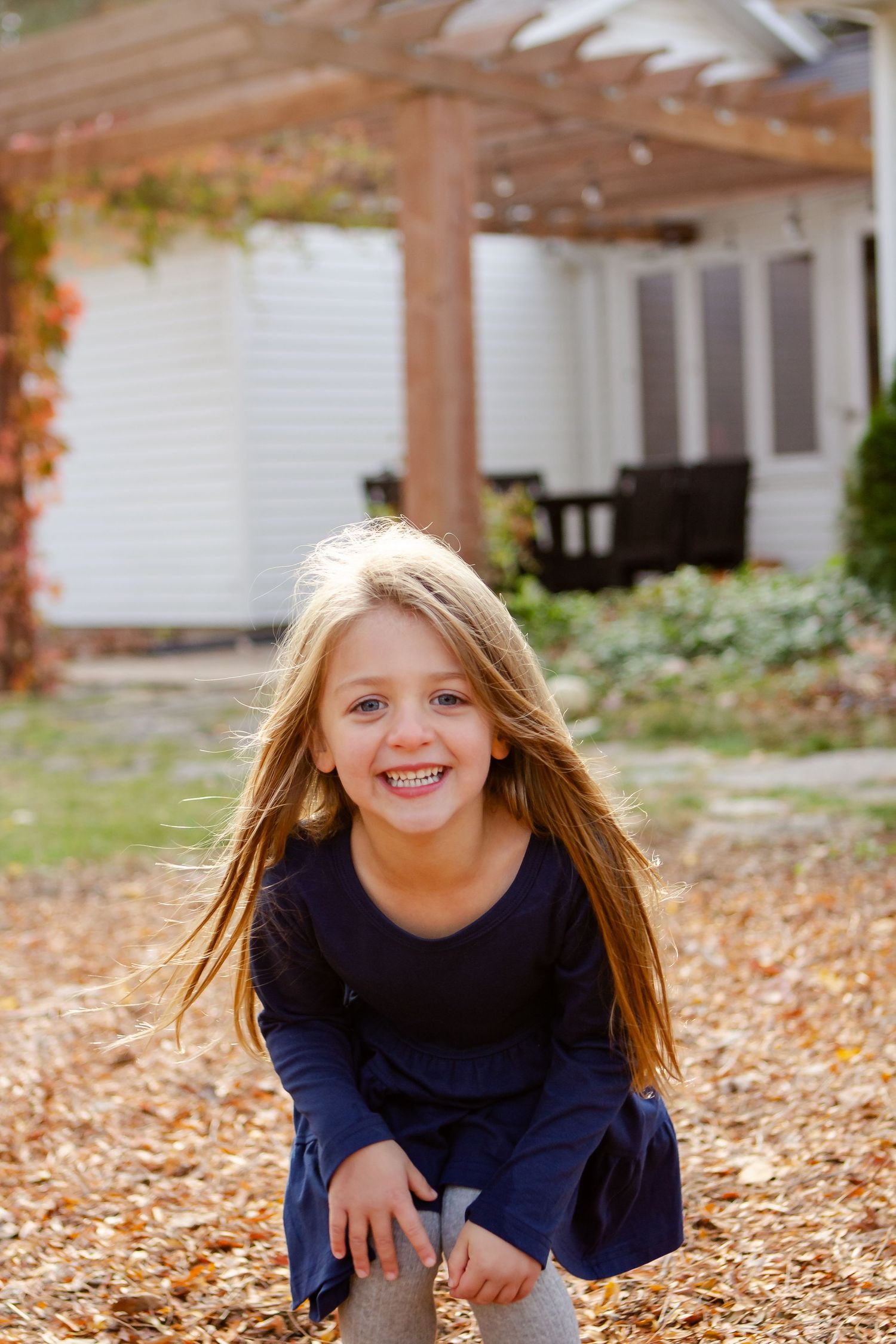 A child plays in fallen autumn leaves under a wooden pergola on a sunny day.