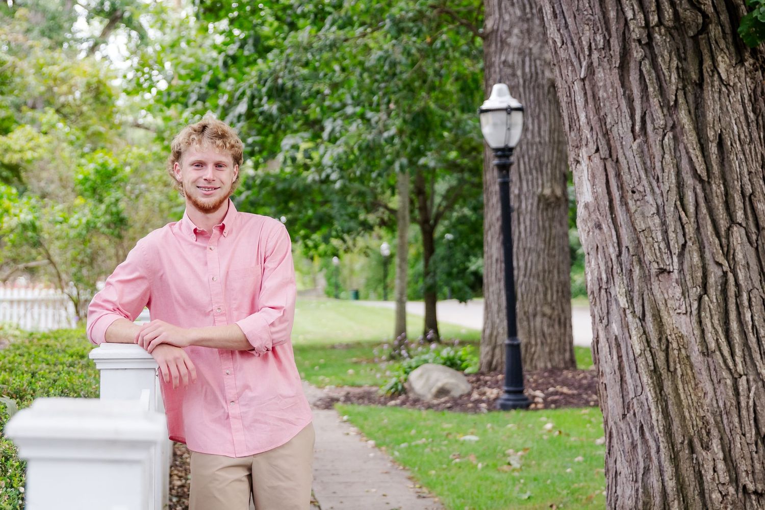 A person in a pink button-down shirt poses outdoors near trees and a white pillar in a natural setting during a Michigan senior session