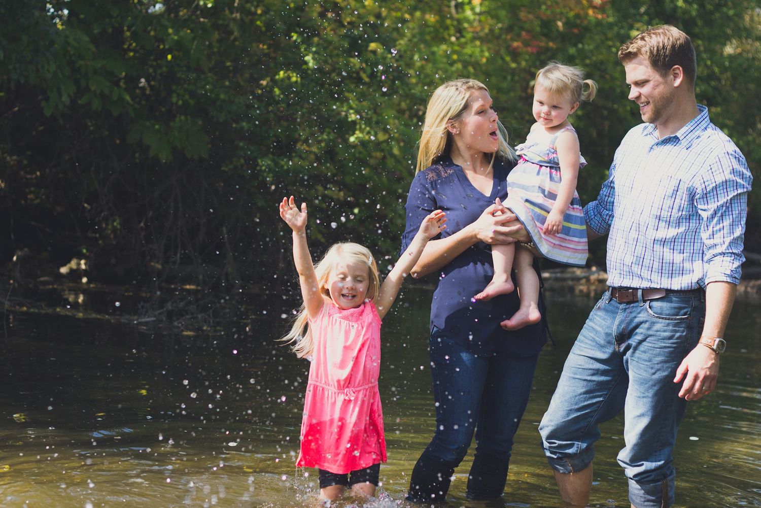 A group enjoys a summer day splashing and playing in a creek or stream with trees in the background during a Delafield photo session
