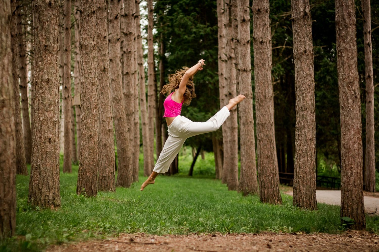 Dancer in pink top and white pants leaps gracefully between rows of tall pine trees.