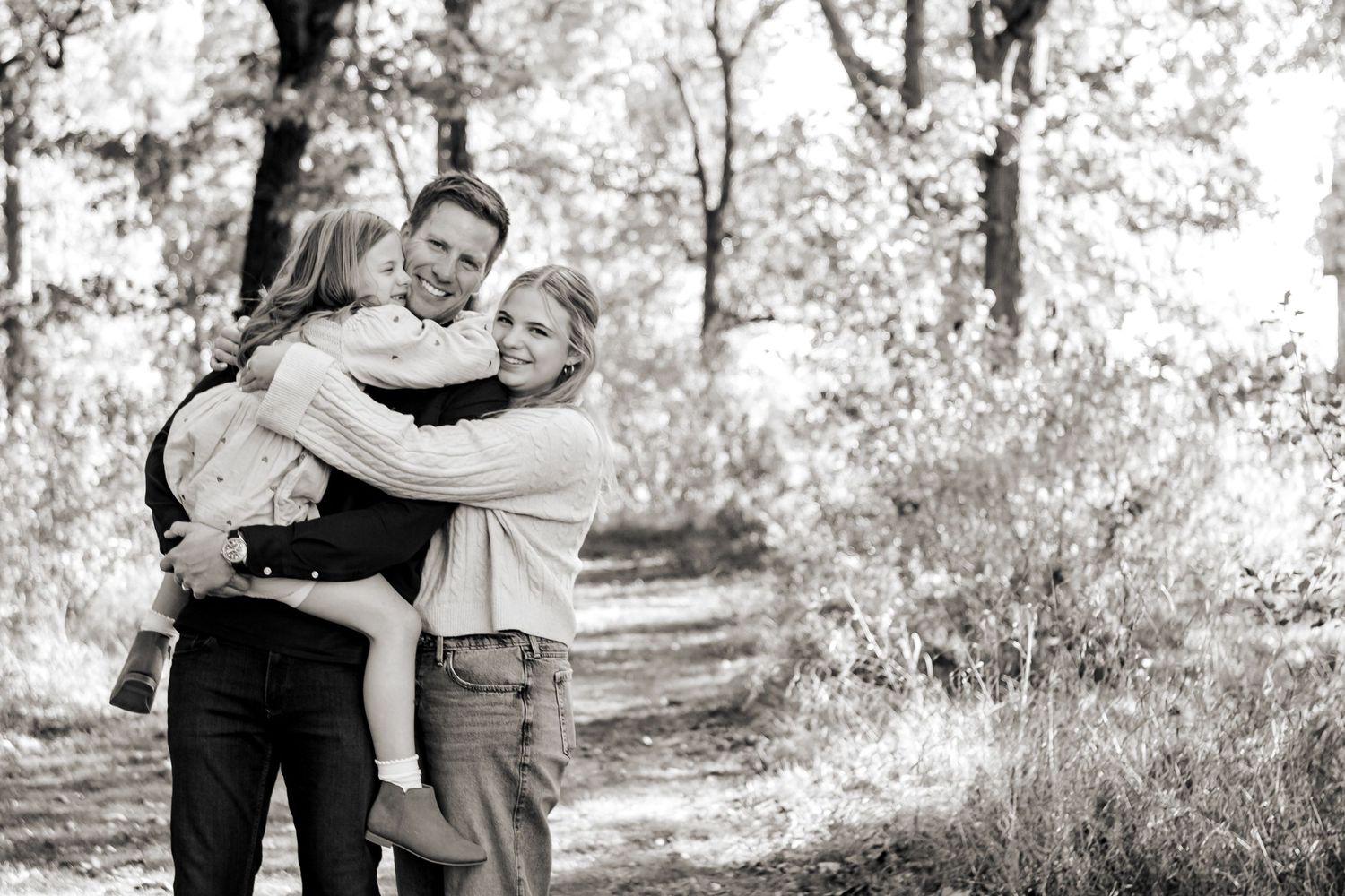 A tender embrace between two people on a sunlit forest path captured in black and white photography at a Lake Country Family session