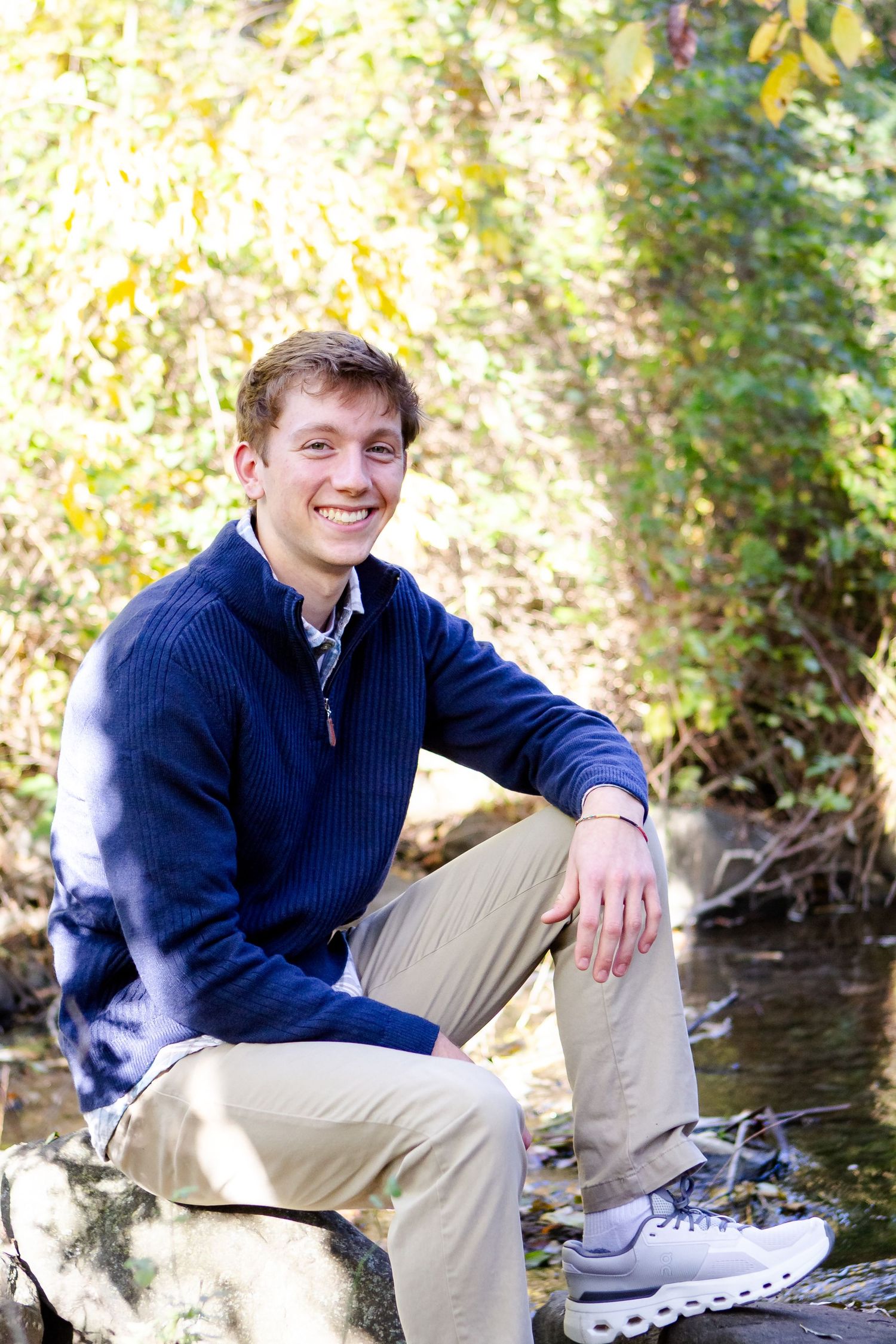 Someone in a navy sweater sits casually on rocks by a stream in an outdoor autumn setting during his Hartland senior session