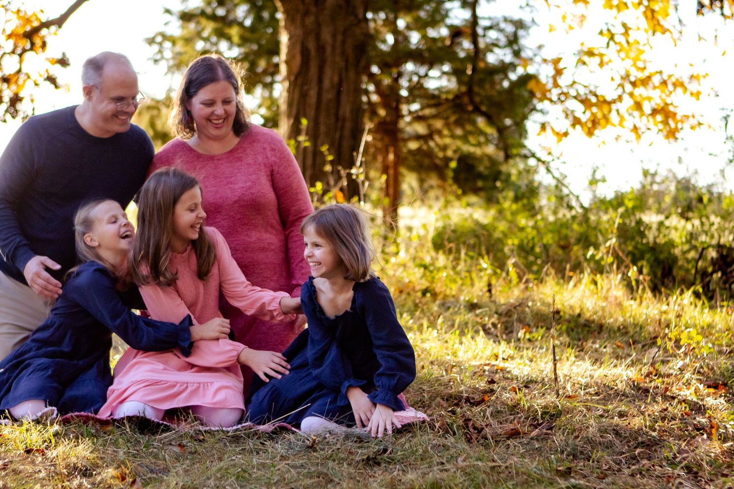 A family sits together outdoors during golden hour in a natural setting with sunlit trees in the background at an Oconomowoc family session
