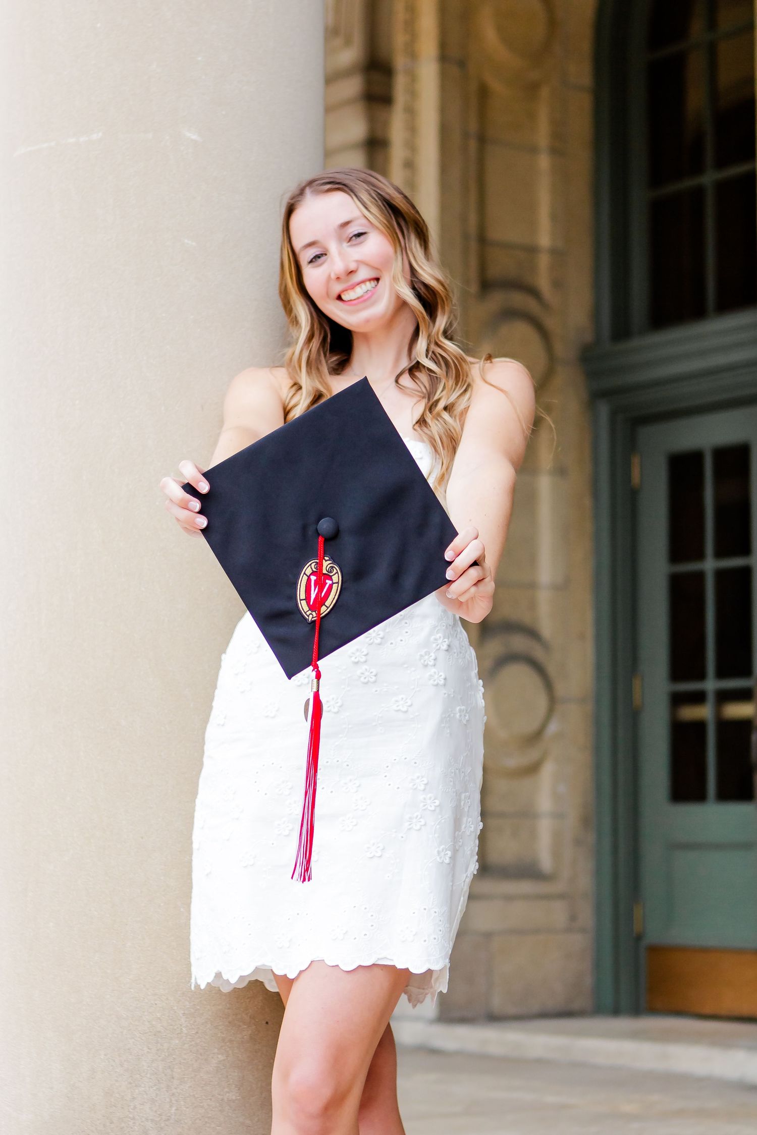 Graduate in white dress and black gown poses against classical architectural columns.