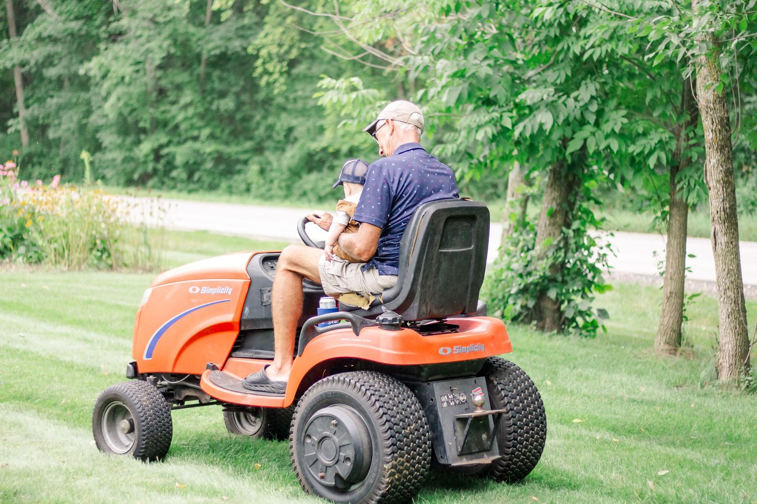 A person operates an orange riding lawn mower in a grassy backyard surrounded by trees at Delafield home session