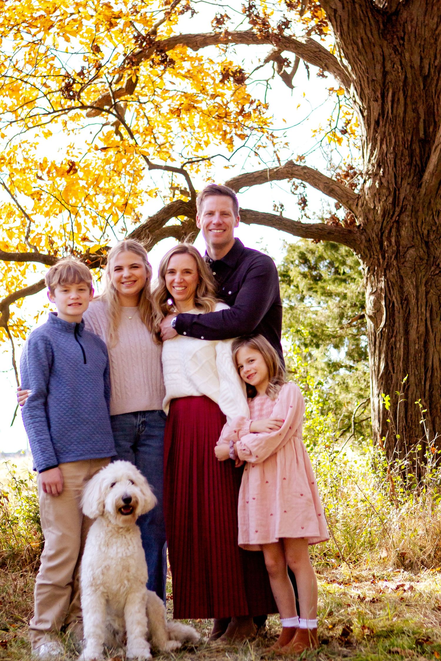 Family poses together with their fluffy white dog under golden autumn trees during outdoor photo session at an Oconomowoc family session