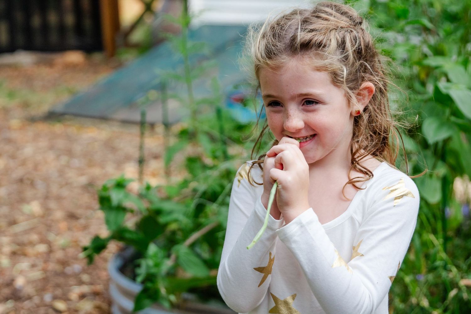 Young child with curly hair wearing a white long sleeve shirt stands in a garden smiling with hand near face at Delafield home session