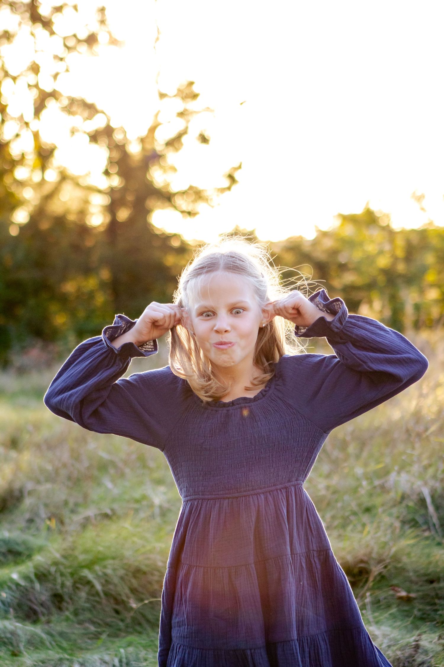 A person in a navy blue dress poses playfully in a field at golden hour with sunlit grass in the background at an Oconomowoc family session