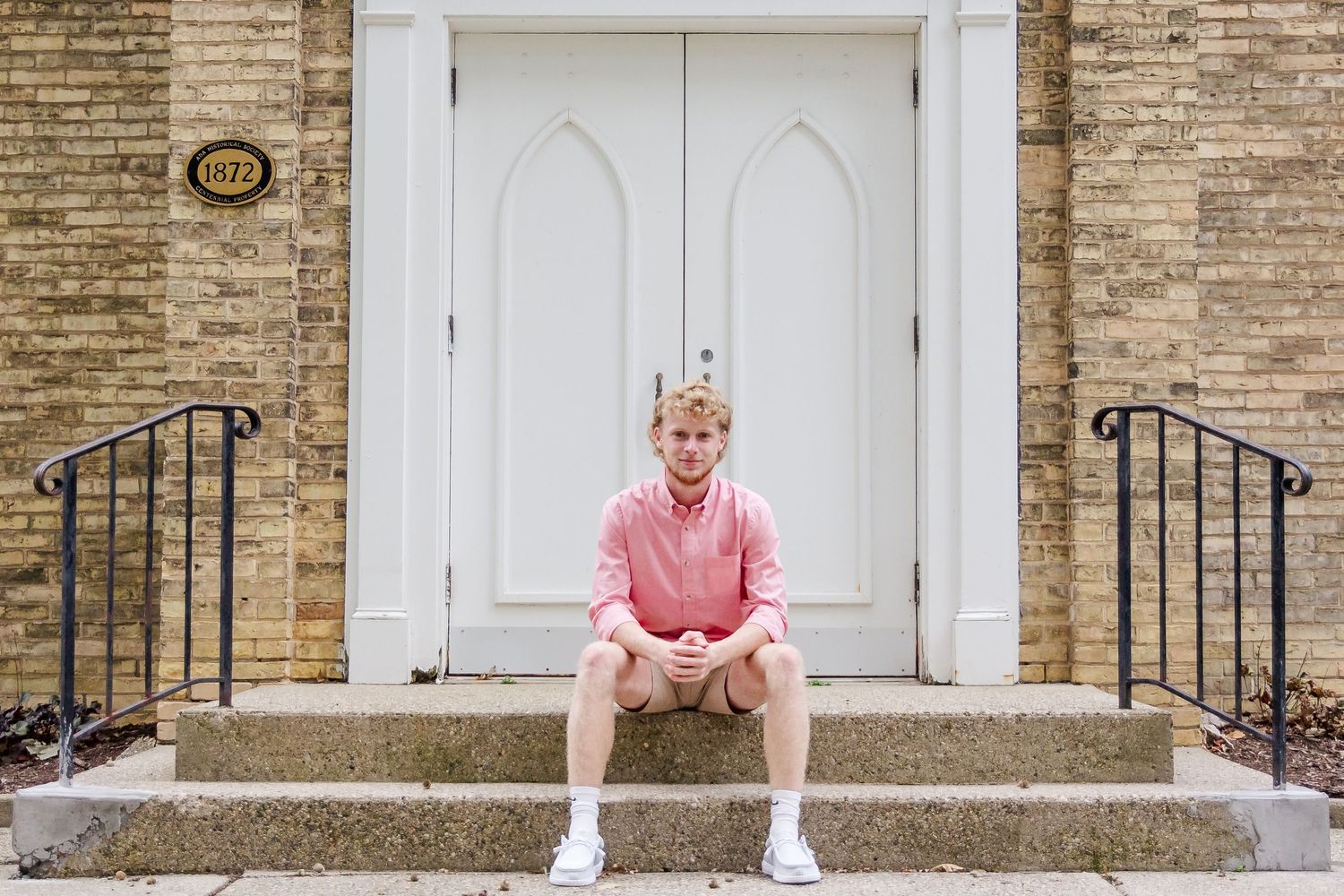 A person in a pink shirt sits on concrete steps in front of a white double door entrance with brick walls during a Michigan senior session