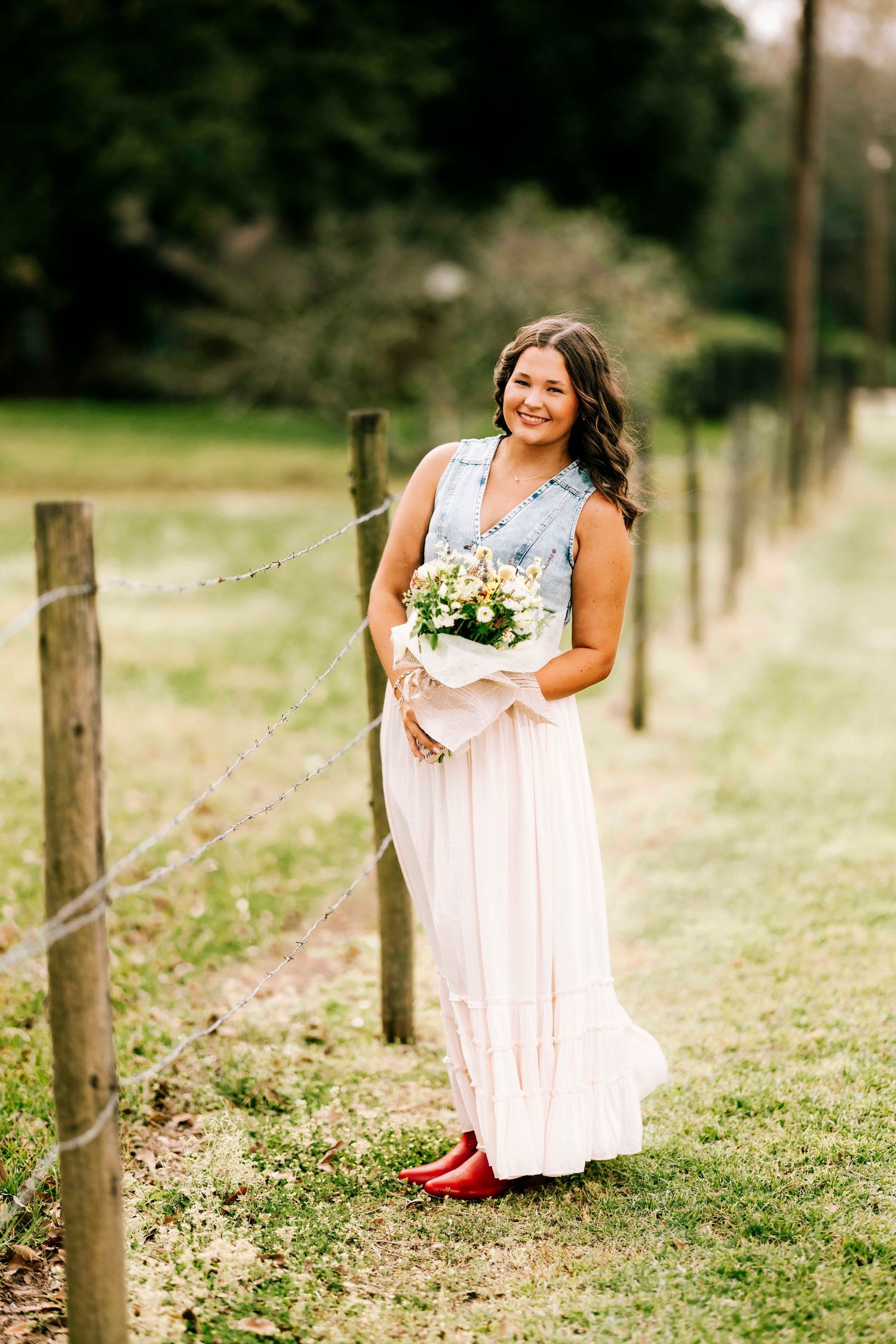 A person in a white maxi dress holding a bouquet stands along a rustic fence in a grassy field.