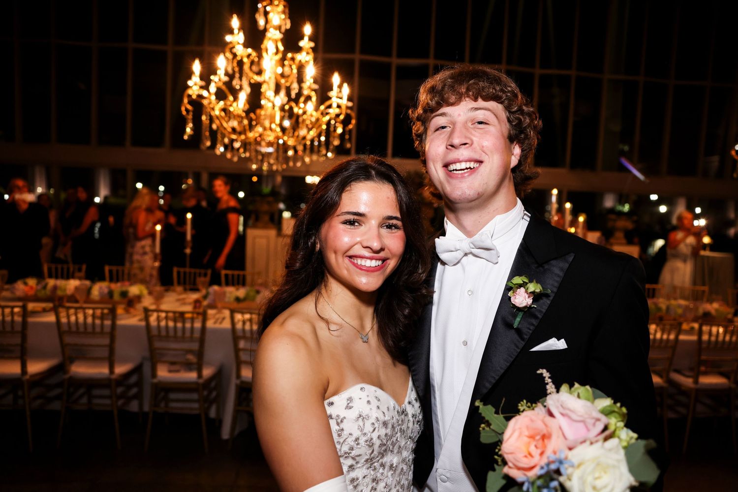 A couple smiles together at their wedding reception with a crystal chandelier glowing overhead.