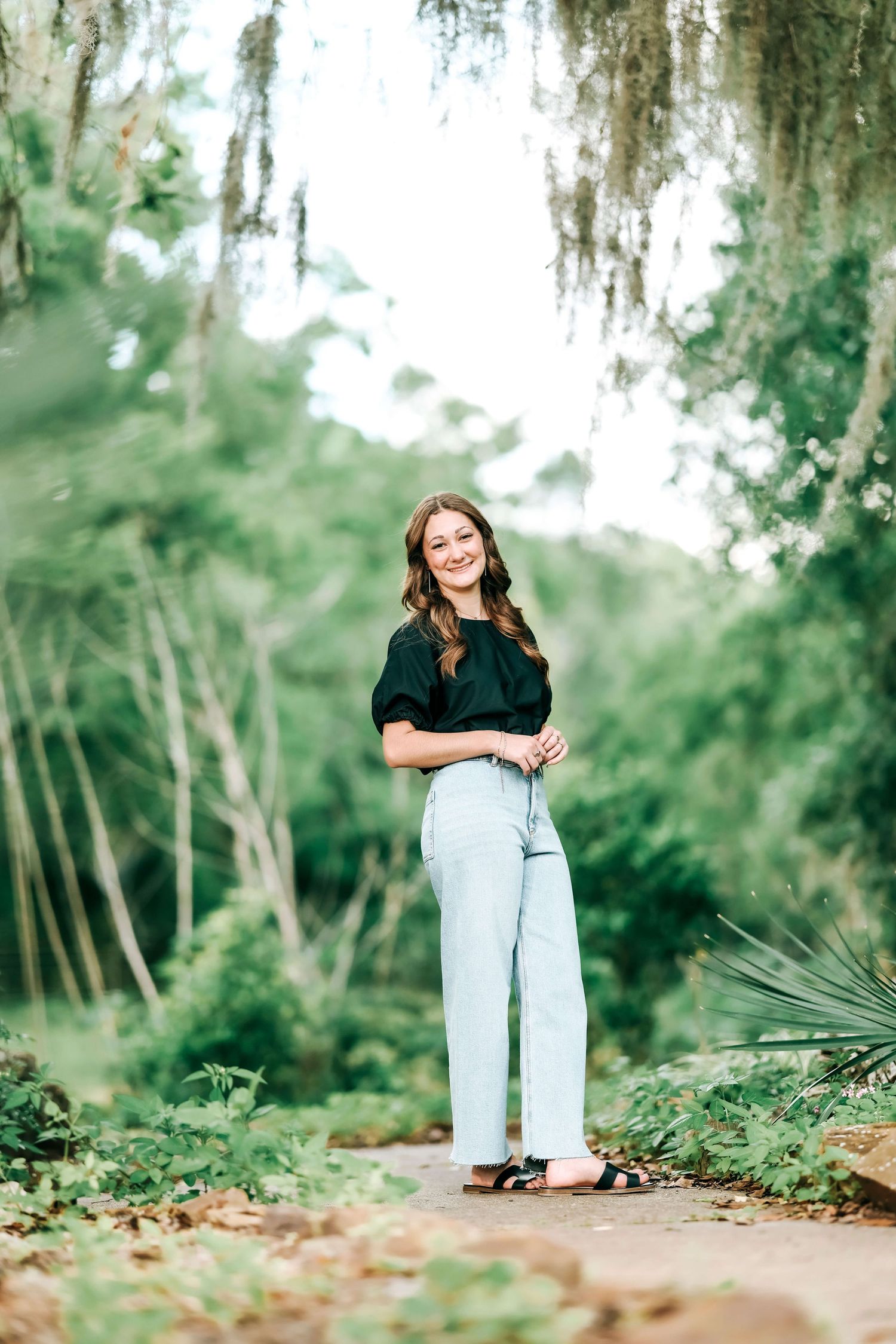 A person in a black top and light jeans poses on a path surrounded by lush greenery.