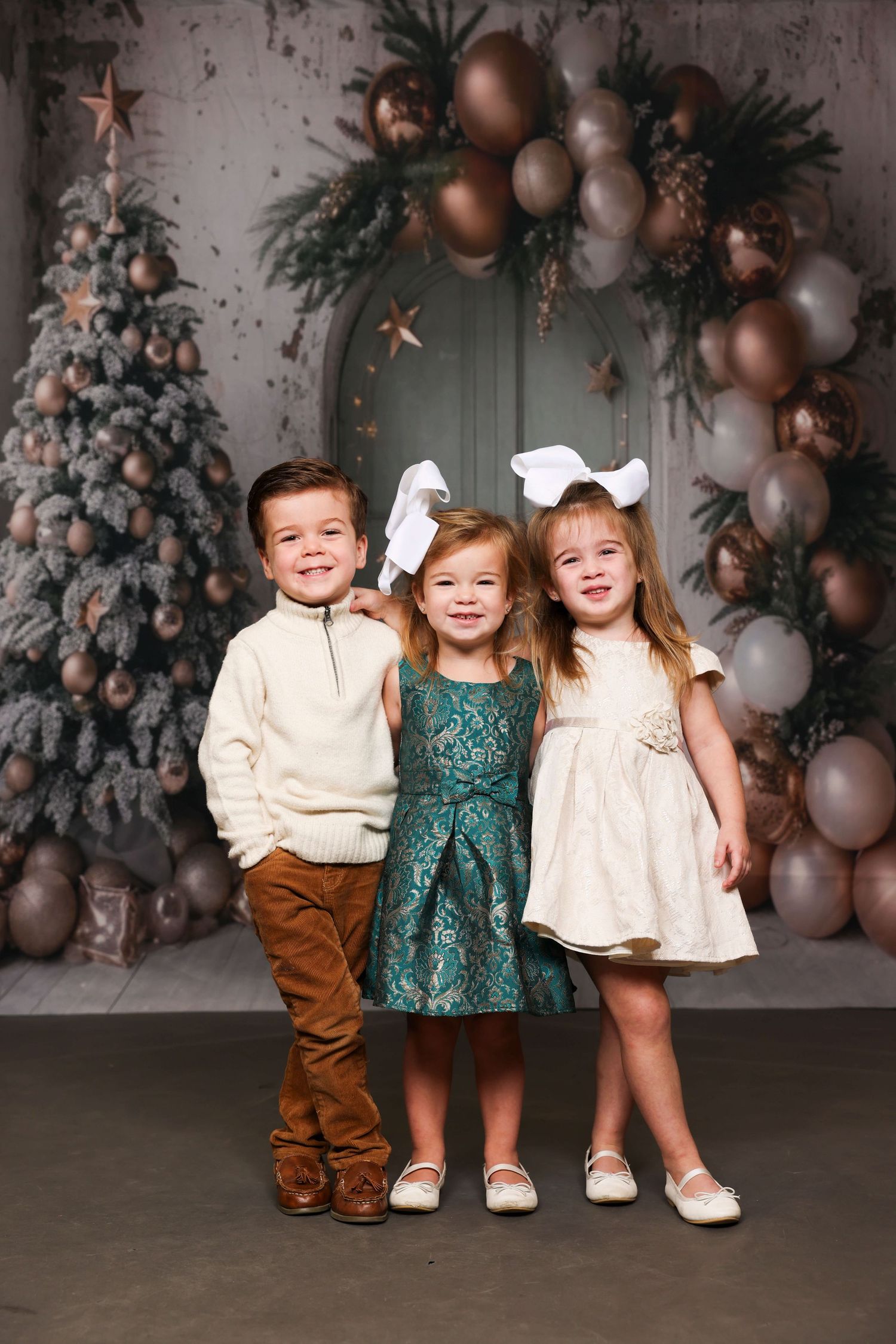 Three young children pose together in front of a decorated Christmas backdrop with balloons and trees.