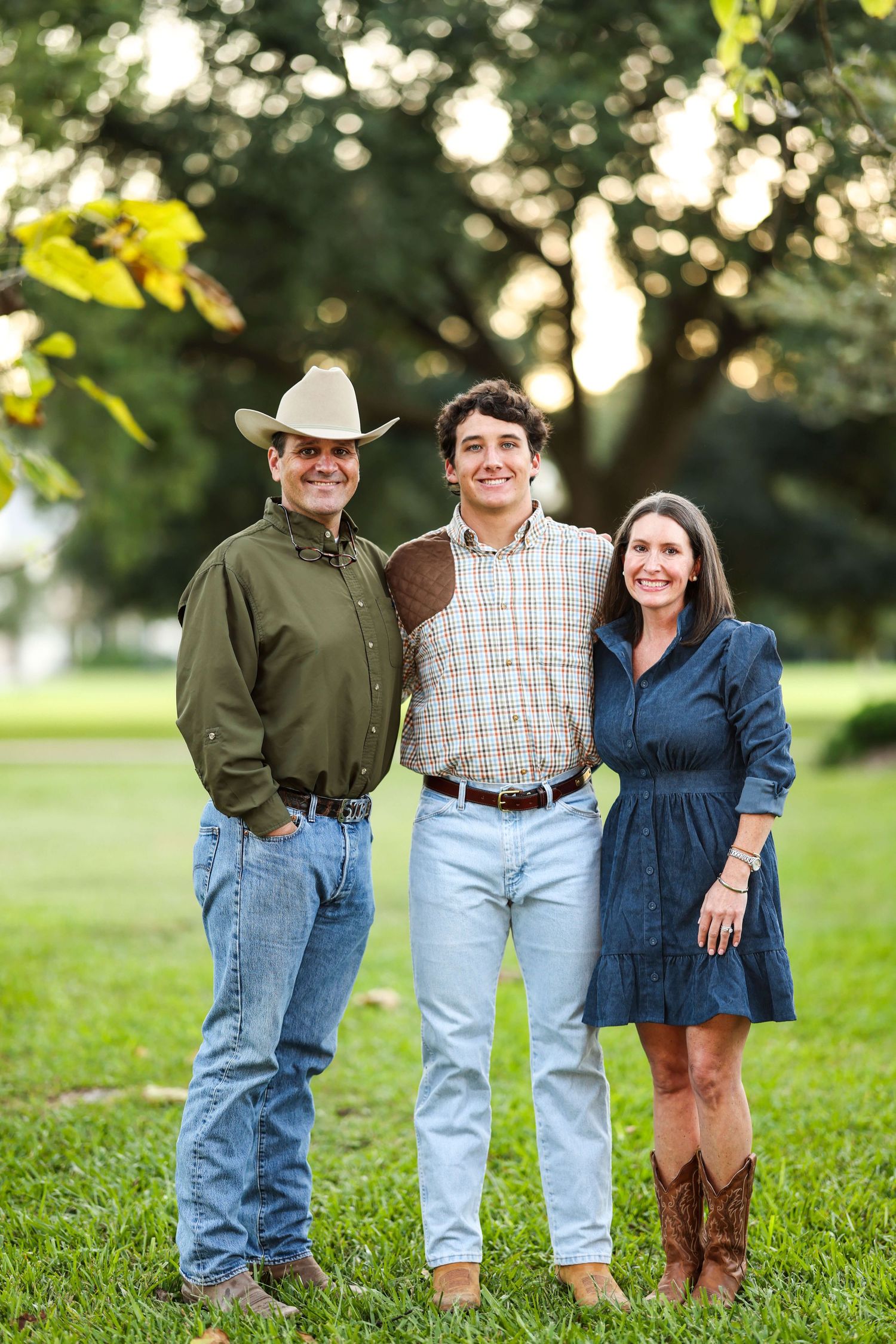 Three people in casual western attire pose together outdoors on grass with trees in the background.