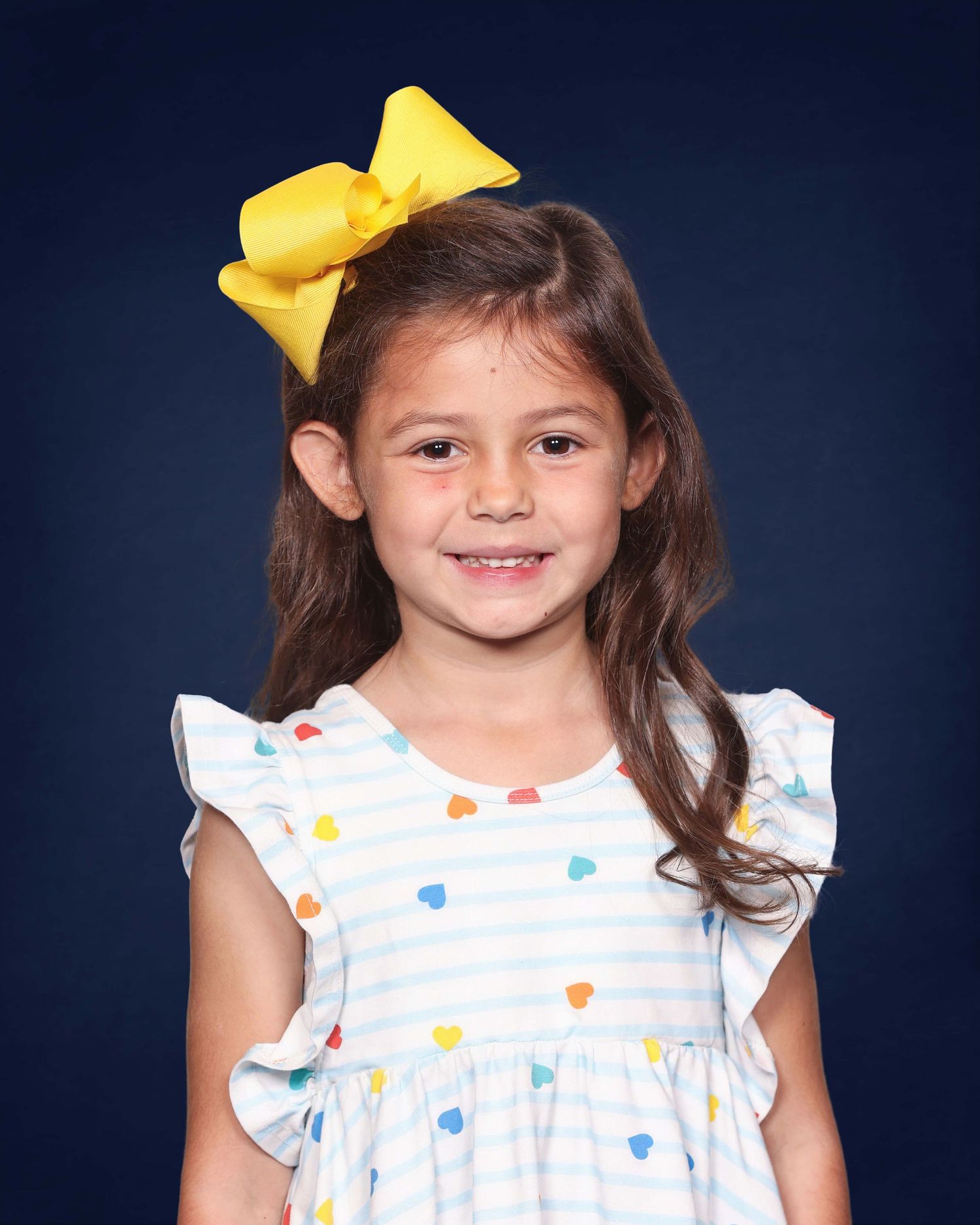 A young student in a white dress with colorful heart patterns and a bright yellow bow against a navy background.