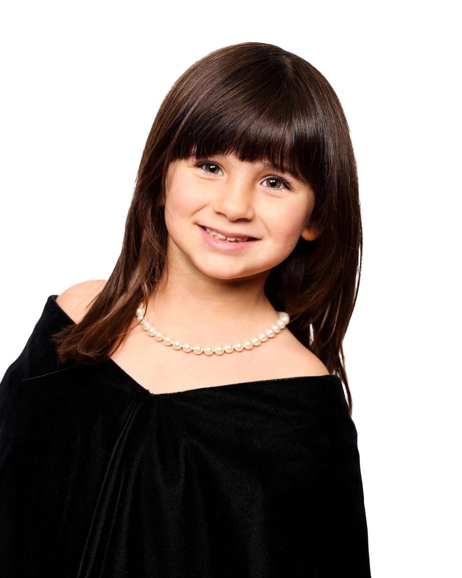 Studio portrait of a young child with dark bangs wearing a black top and pearl necklace against a dark background.