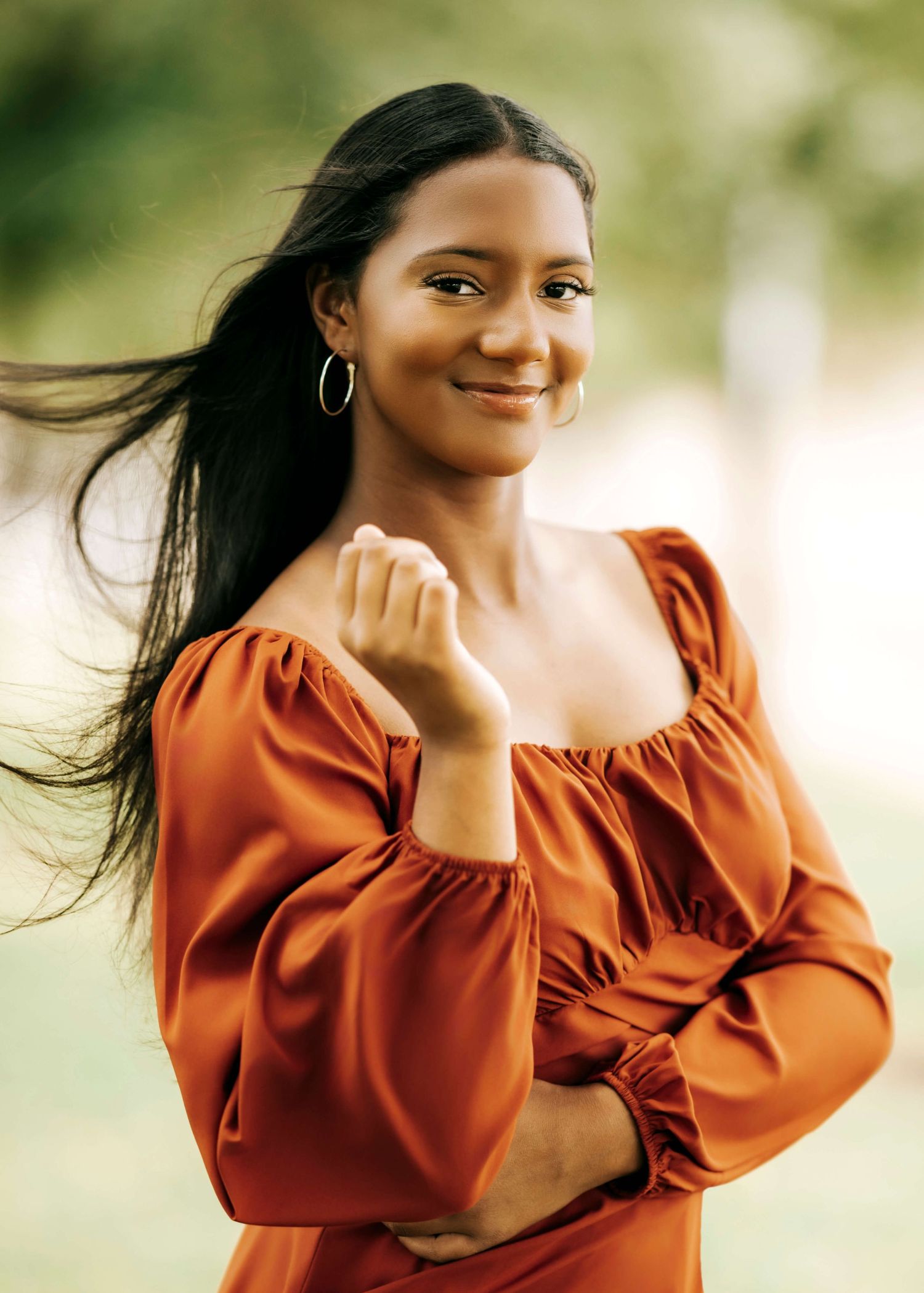 Woman in rust-colored top poses with flowing hair against soft natural background.