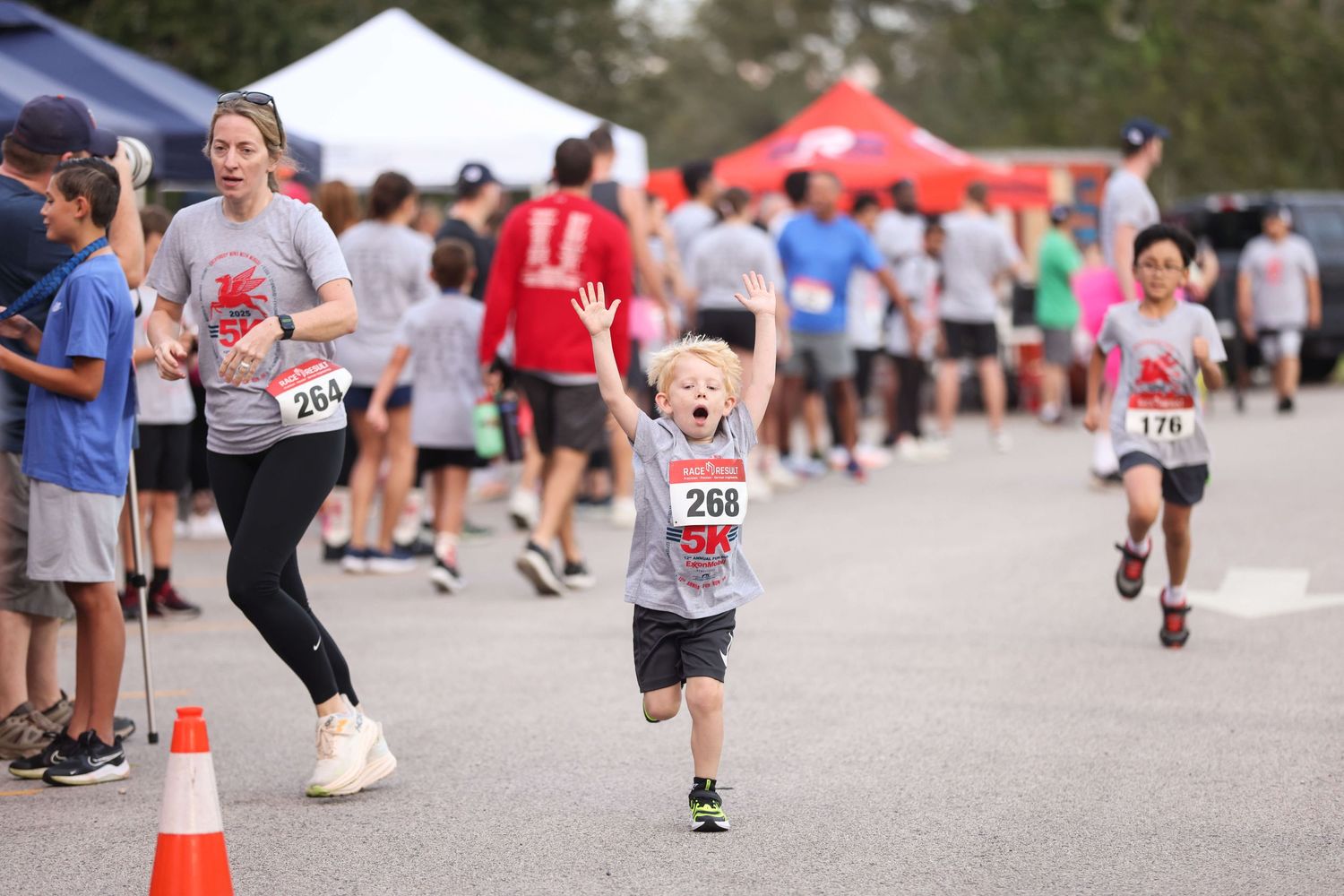 Young participant runs enthusiastically during a community race event with spectators and tents in the background.