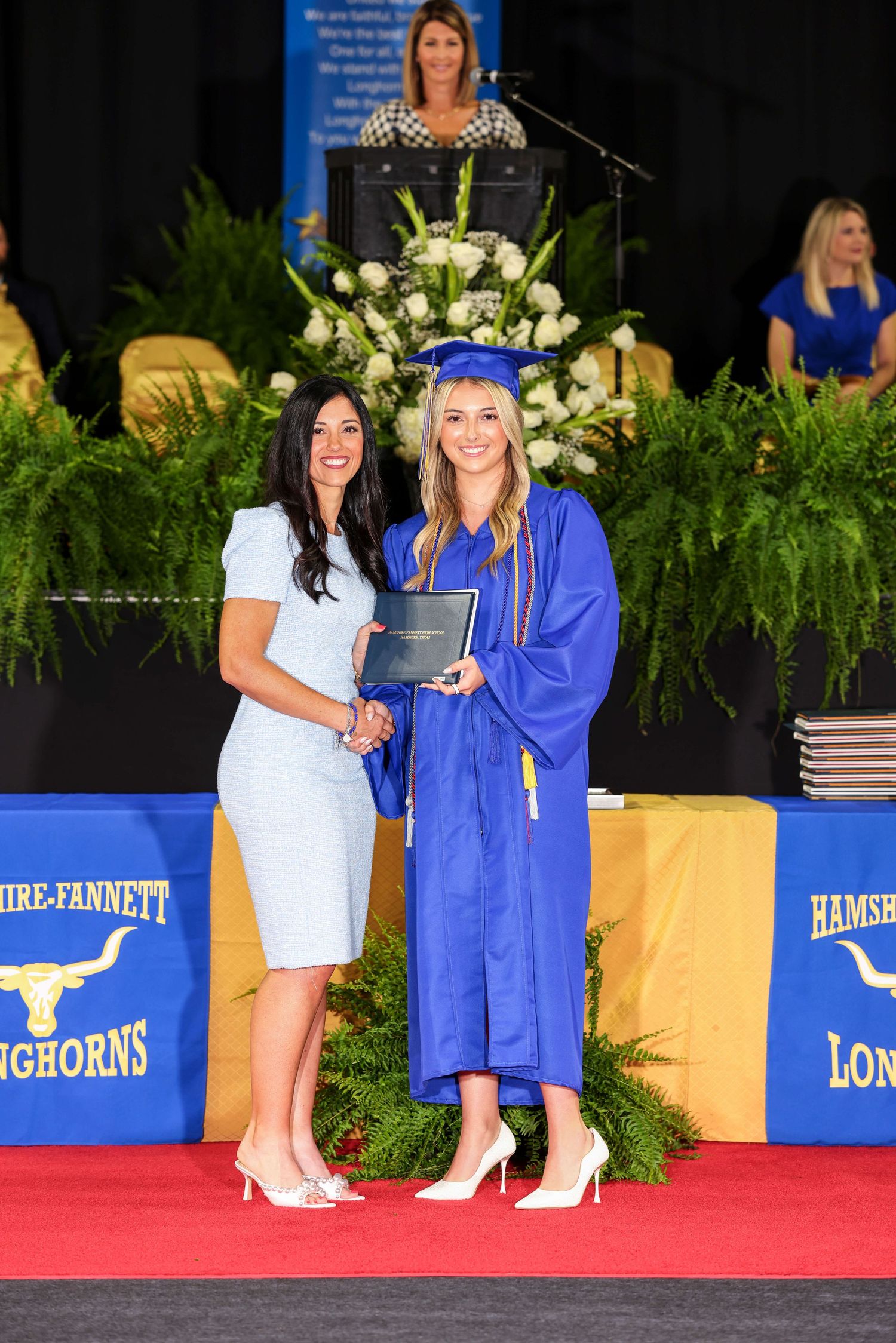 Students in blue graduation gowns and white dresses pose for photos during commencement ceremony on red carpet stage.