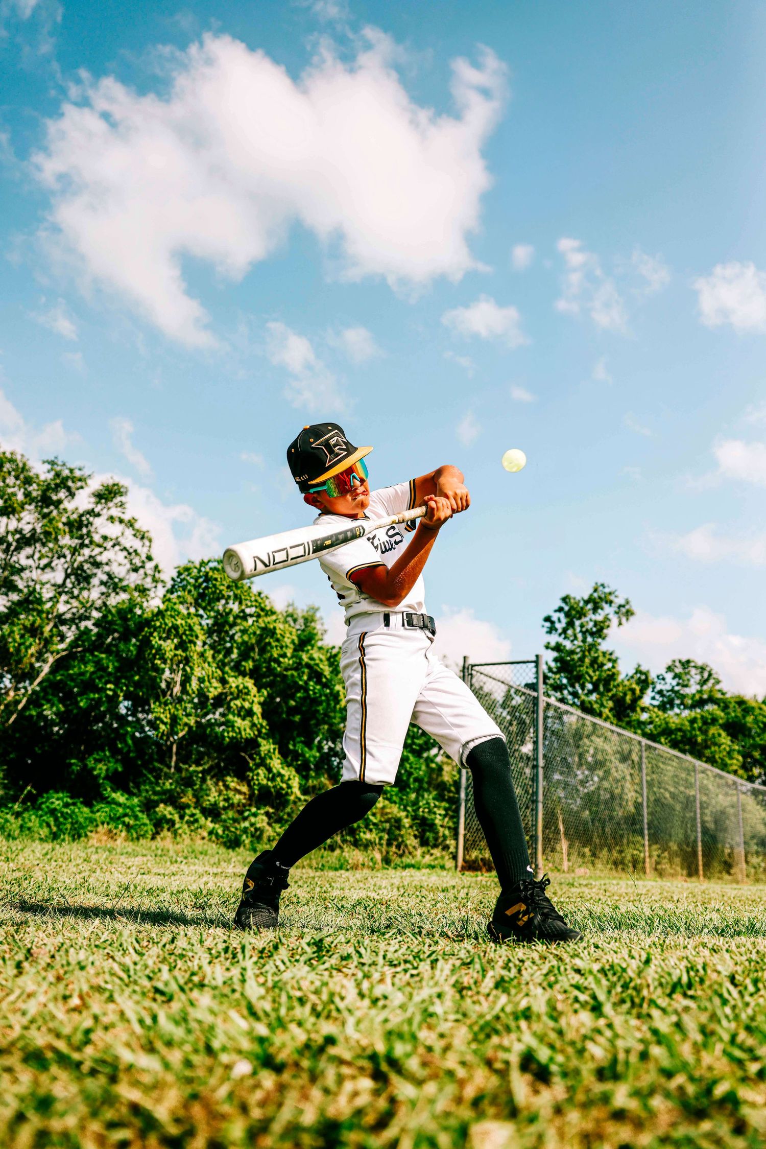 Baseball player practices batting form on a sunny field with blue sky and green grass.