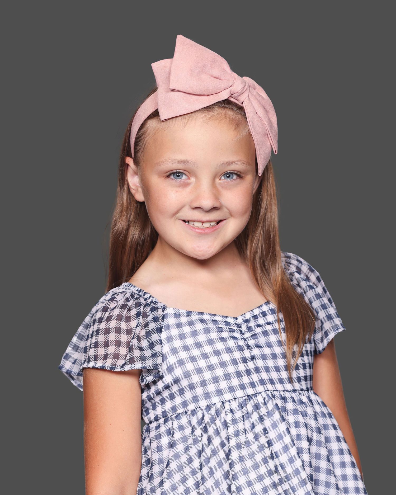 Young child wearing a blue and white gingham dress with pink bow headband against grey background.