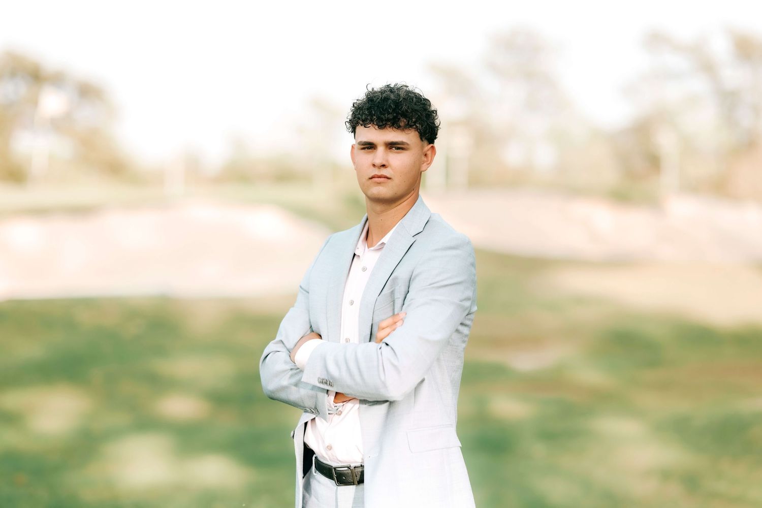 A person in a light gray blazer stands confidently with arms crossed in an outdoor setting.