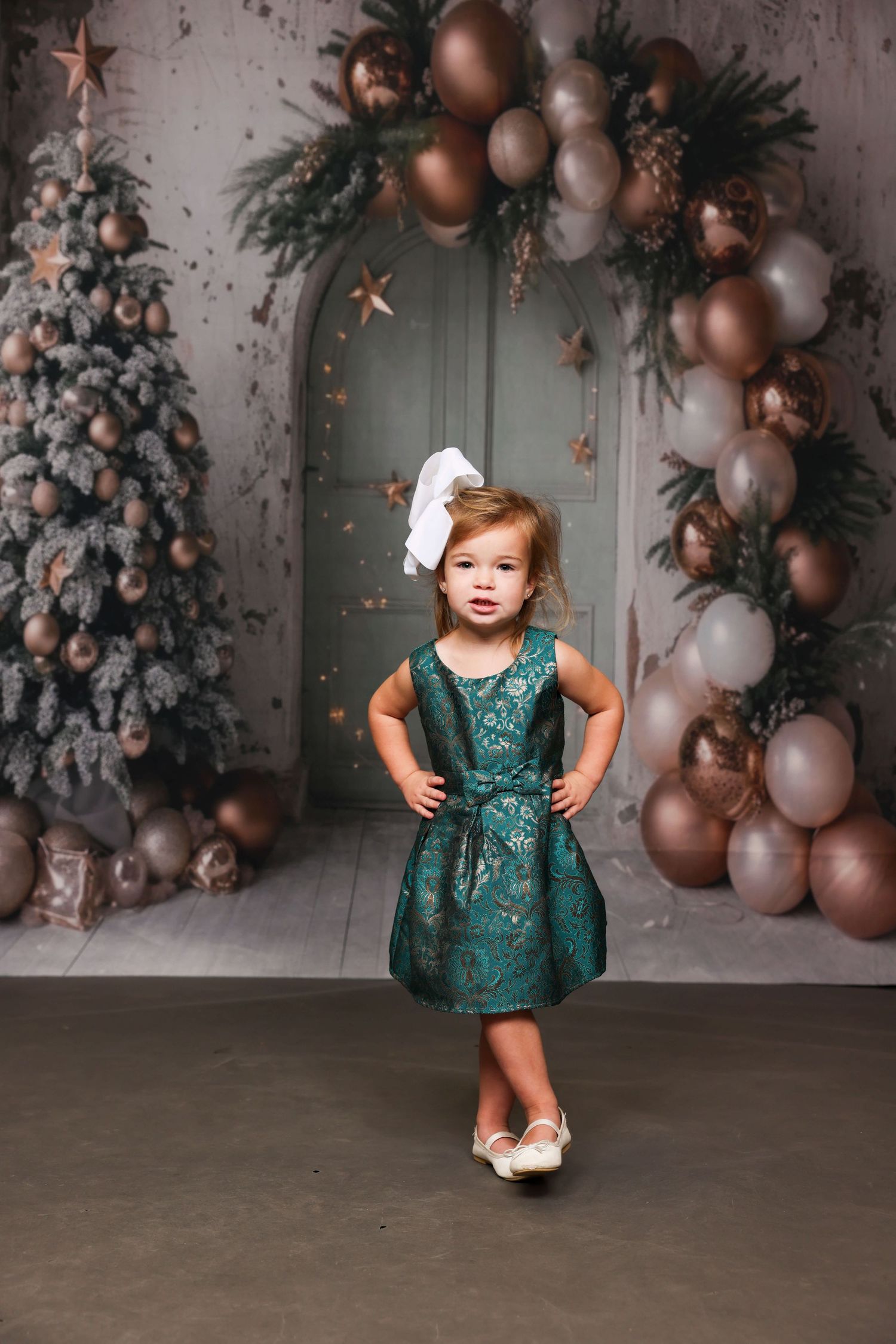 Holiday photo scene with decorated Christmas tree, balloon arch, and sparkly green dress against grey backdrop.