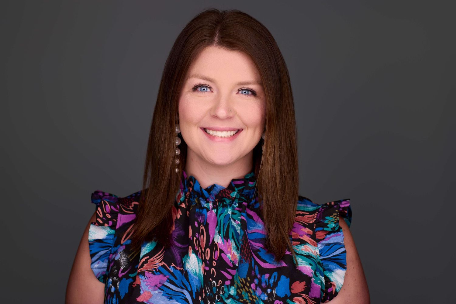 Professional portrait of person wearing colorful ruffled blouse against gray backdrop.