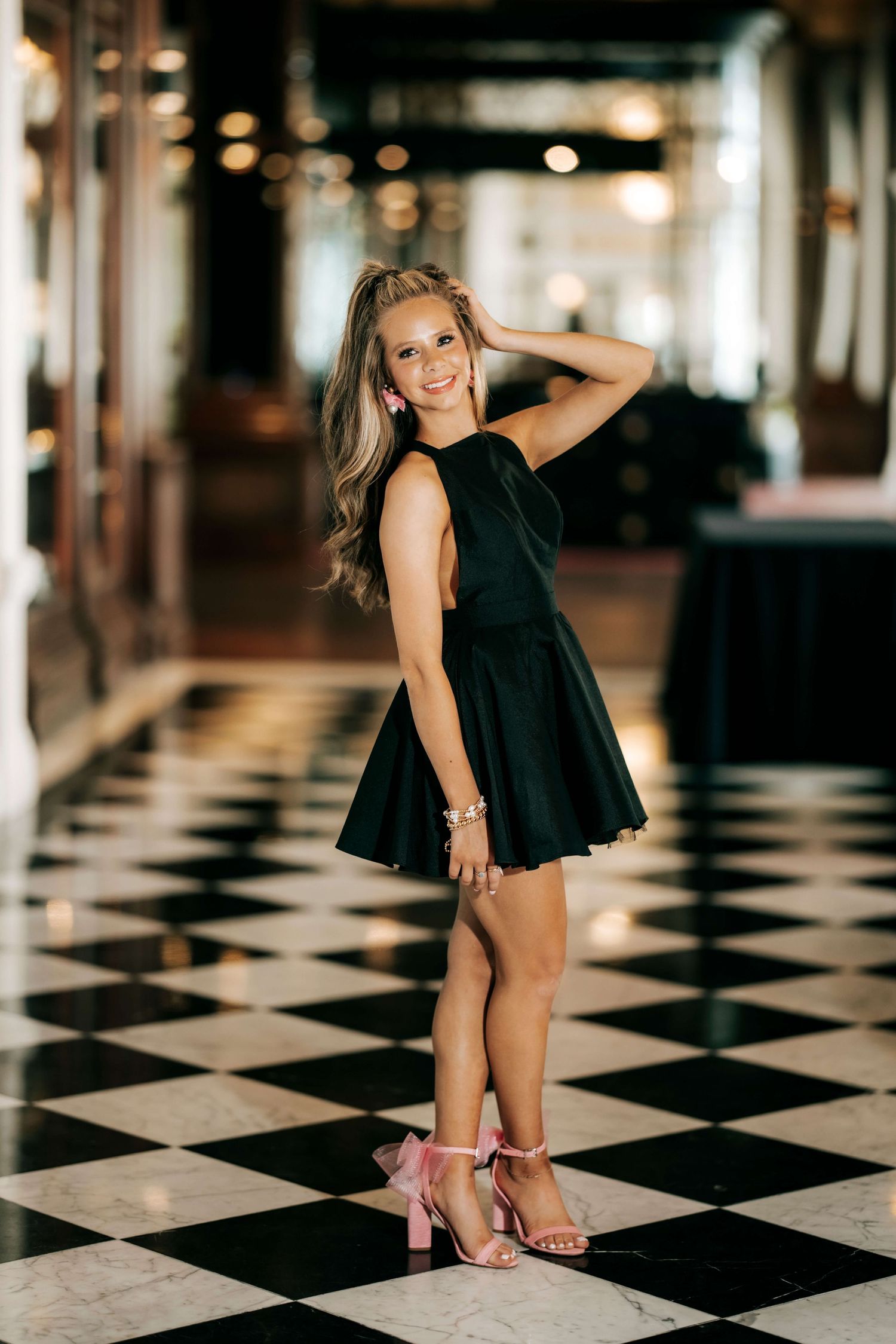 A person in a black dress poses on a checkered floor with pink heels.