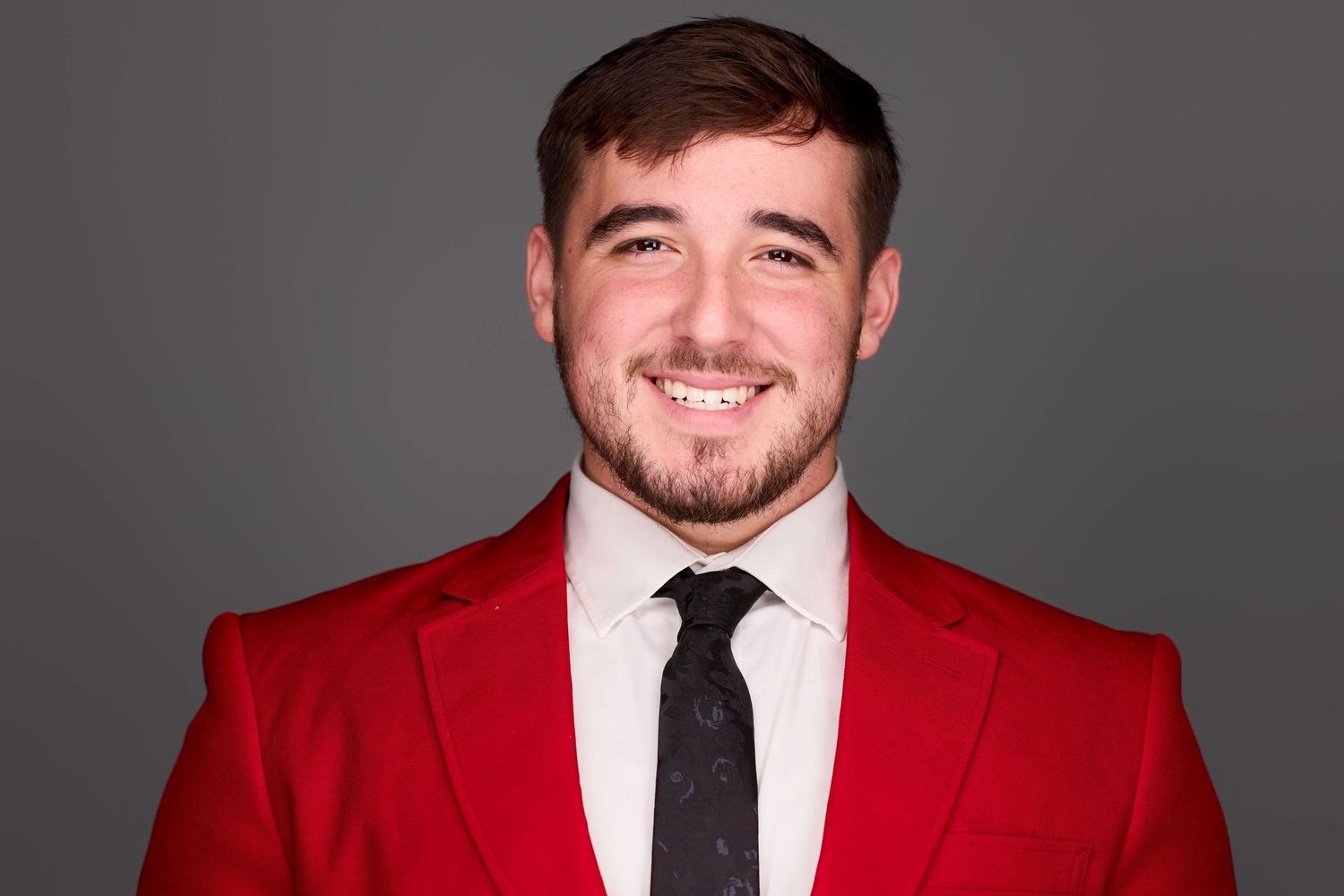 Professional headshot of person in red suit jacket and black tie smiling at camera.