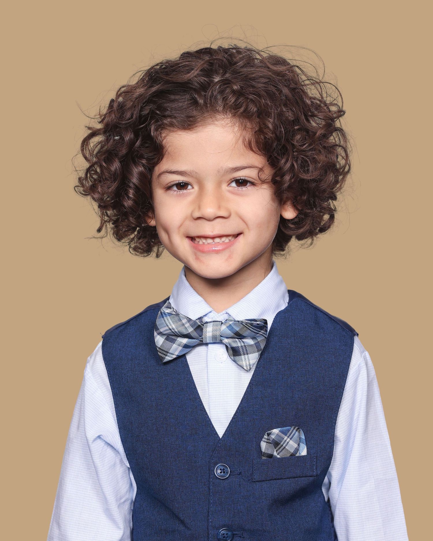 A child in formal attire wearing a navy vest, plaid bowtie and white dress shirt against a tan backdrop.