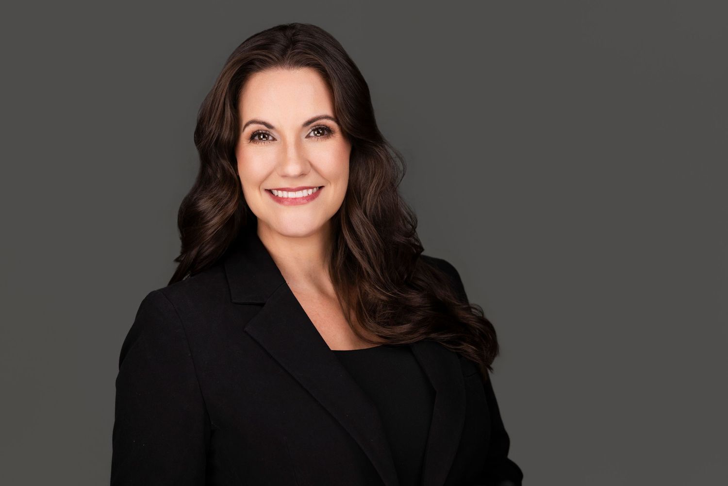 Professional headshot portrait of someone with long dark hair wearing a black blazer against a gray background.