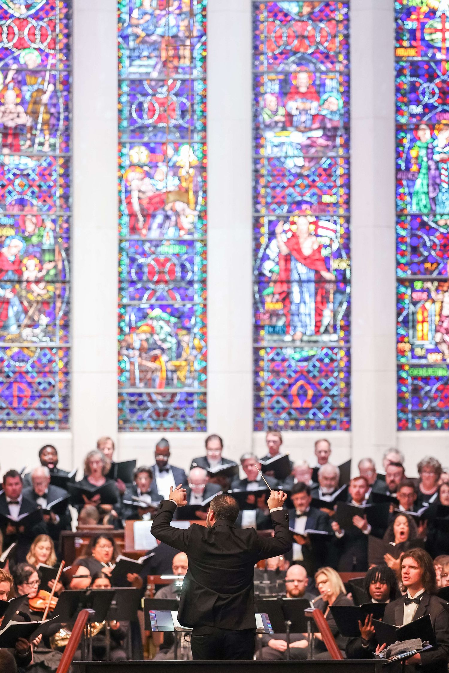 Orchestra and choir perform in front of colorful stained glass windows in a church setting during a concert.