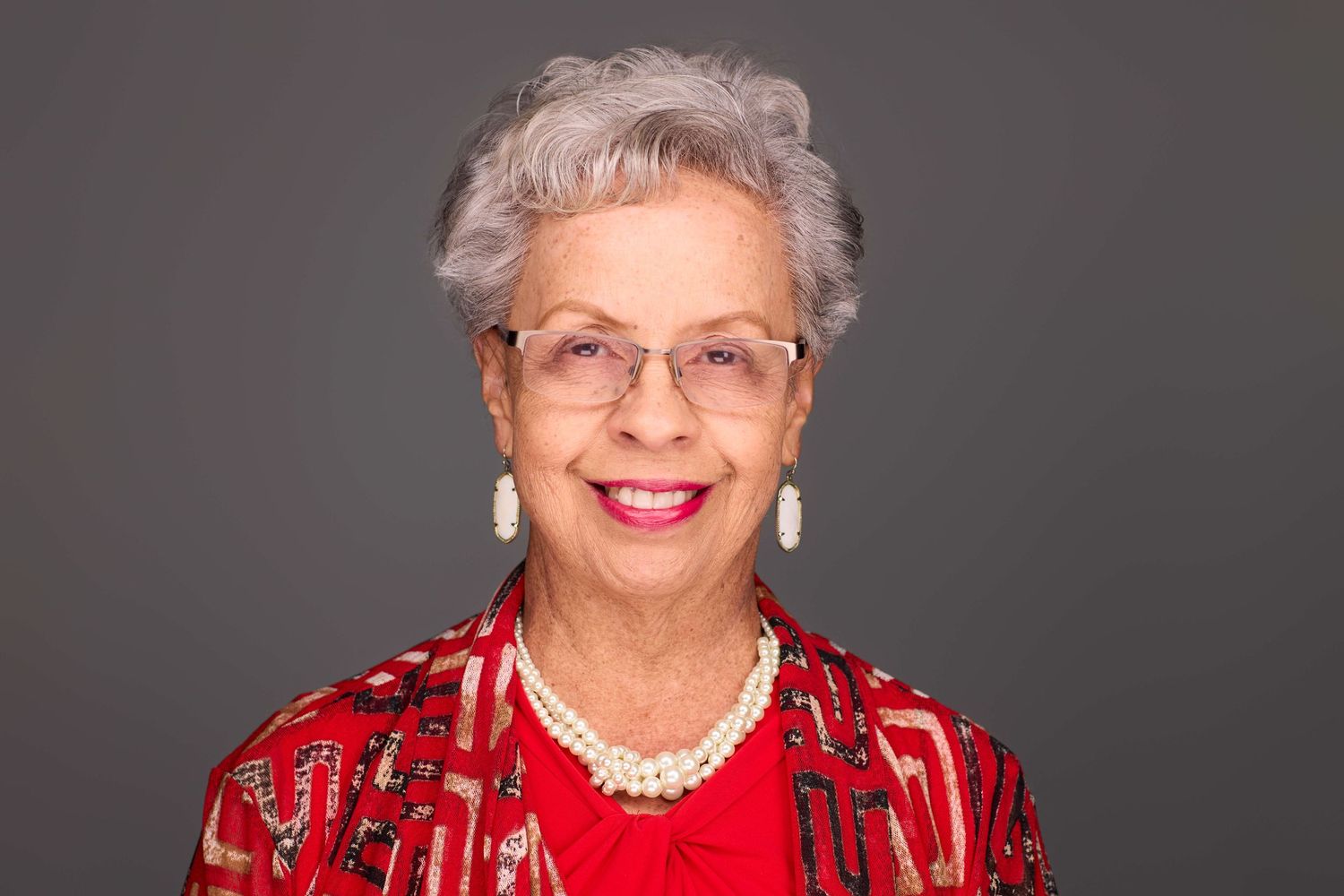 Professional portrait of someone wearing a festive red outfit with pearl necklace against gray backdrop.