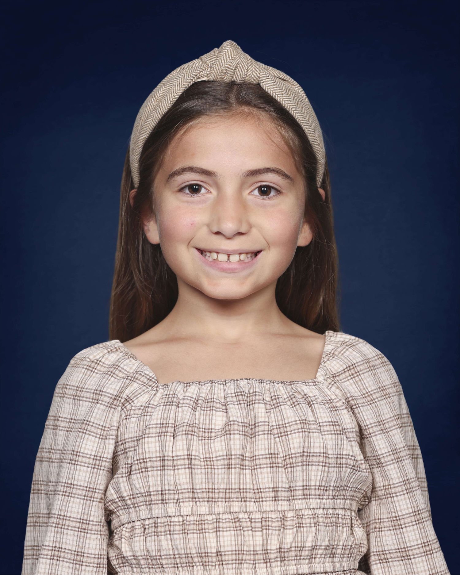 A young person in a plaid dress and beige hat poses against a dark blue background.