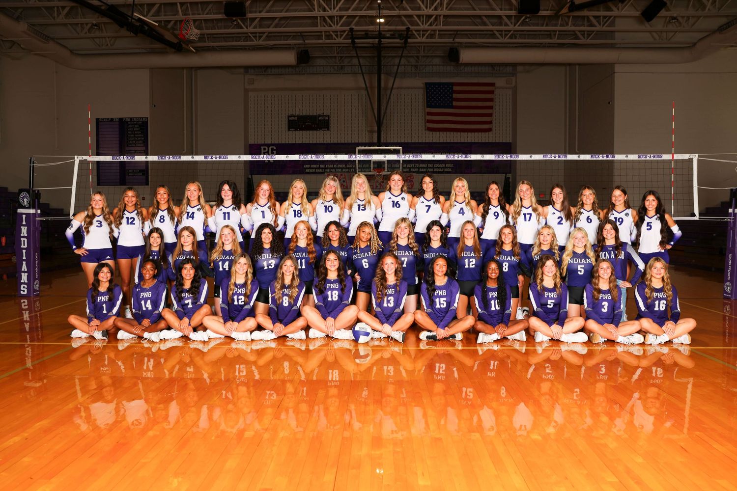 Large volleyball team poses in blue and white uniforms for group photo in gym.