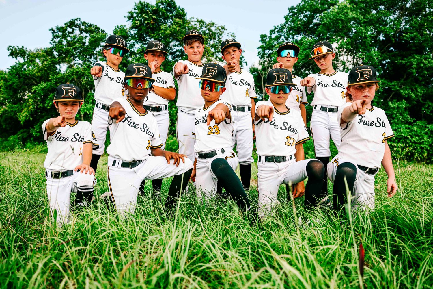 Youth baseball team in white uniforms poses together in grassy field.