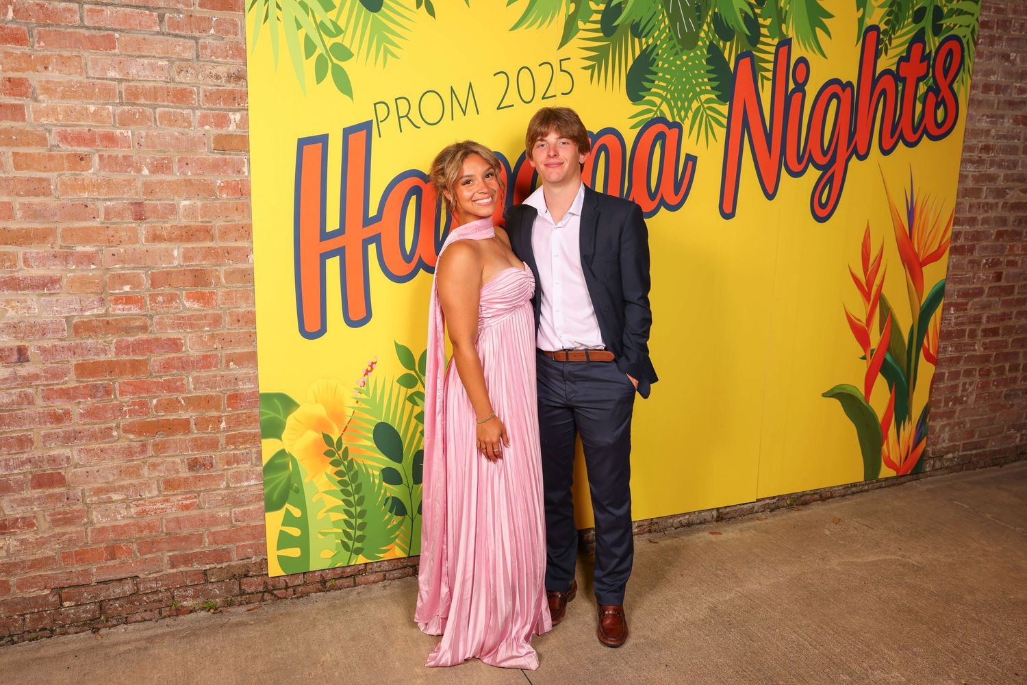A couple poses in front of a bright yellow 'Havana Nights' themed prom backdrop with tropical foliage decorations.