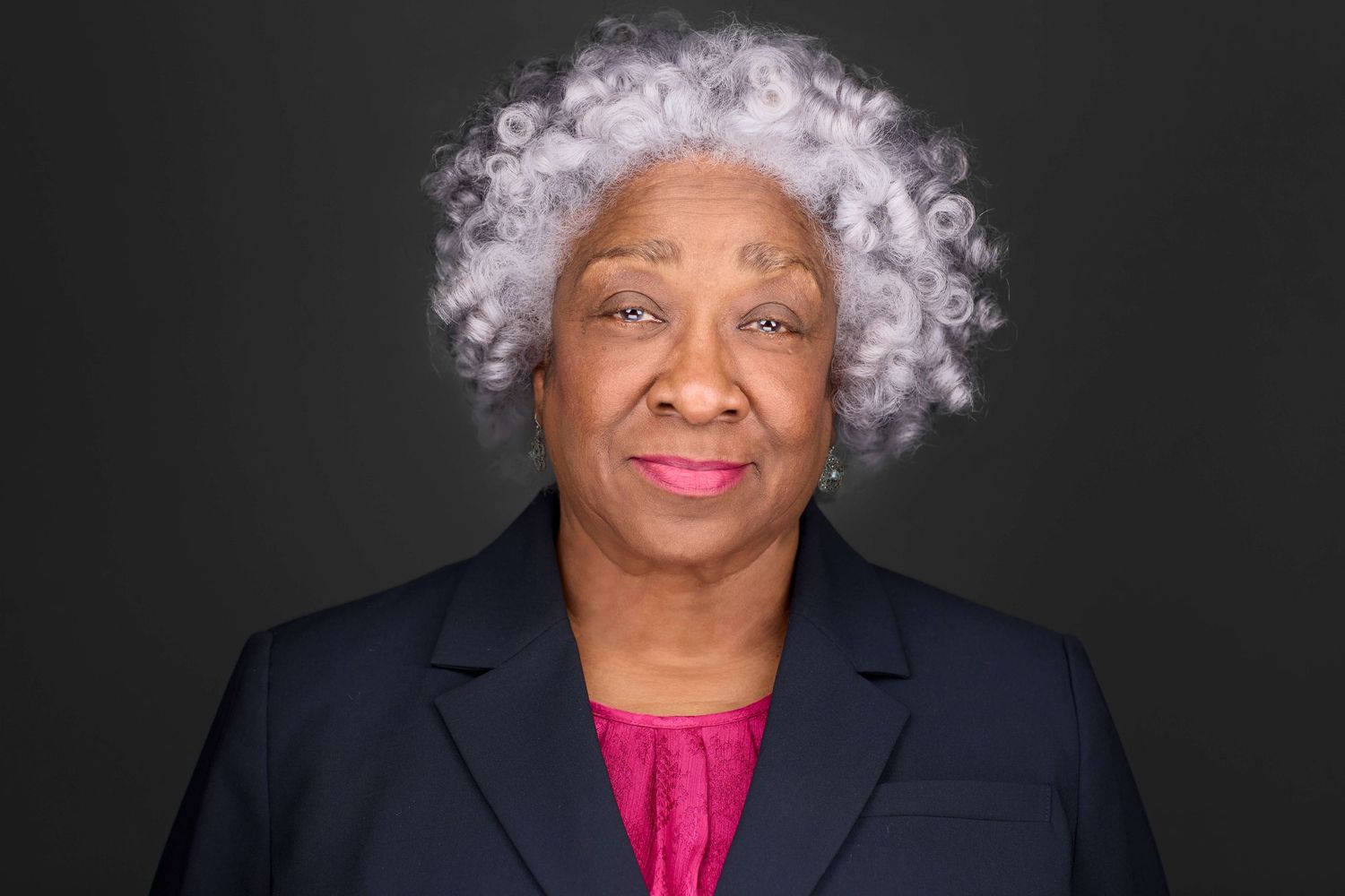 Professional headshot of a person in a navy blazer and pink top against dark background.