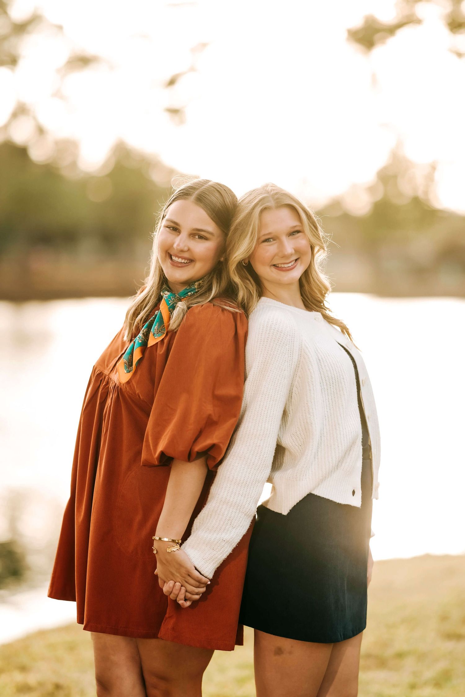 Two friends in casual outfits holding hands and posing back-to-back by a lake at sunset.