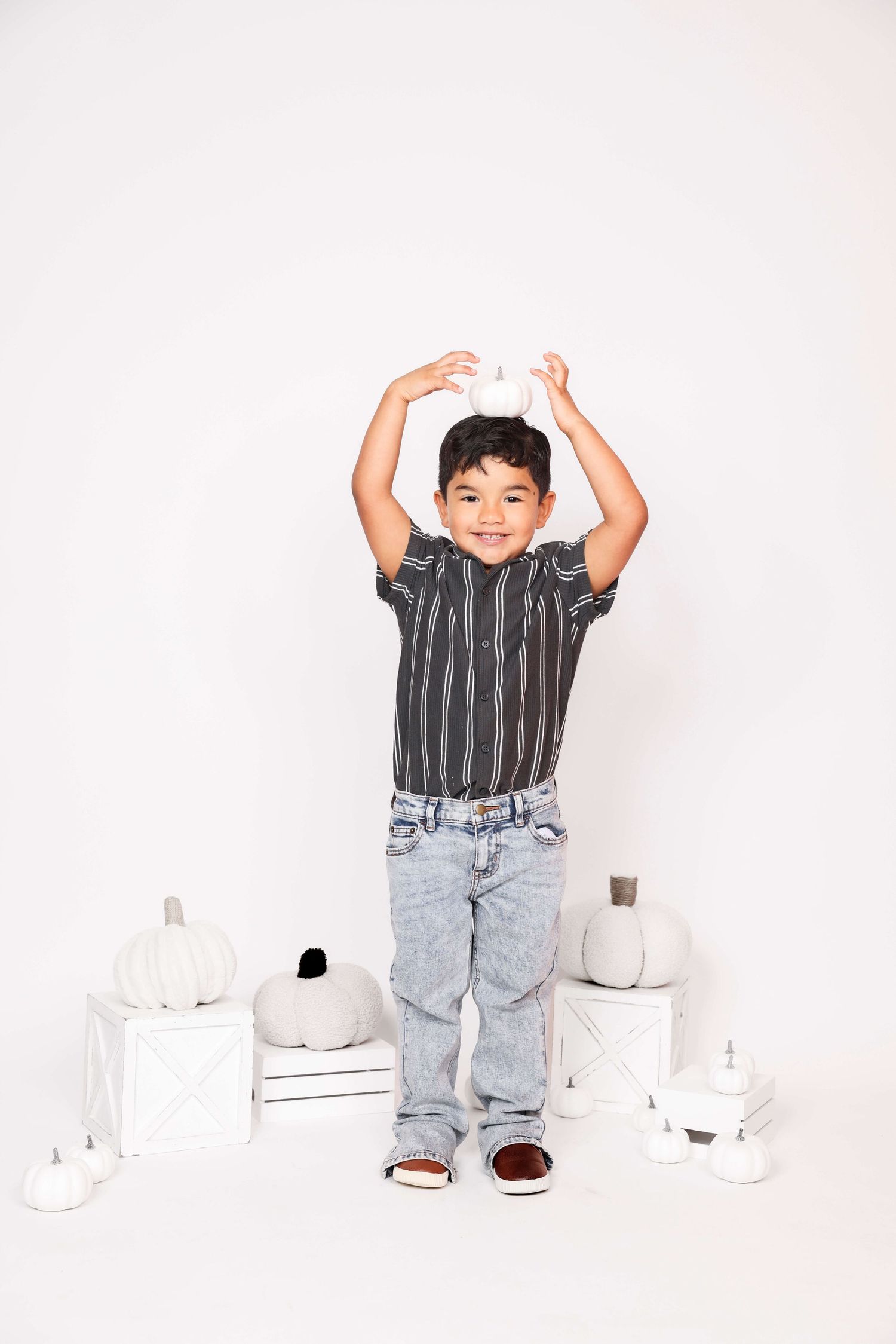 Child in striped shirt and jeans poses playfully with white pumpkins on light background.