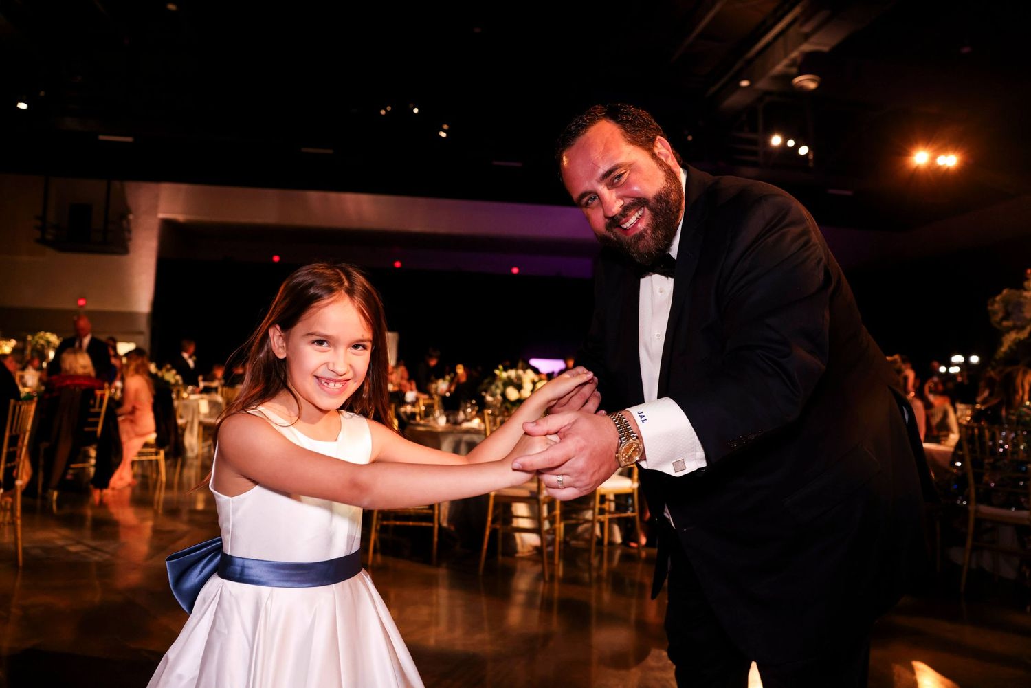 A couple dances together at an elegant evening reception with warm lighting and other guests in the background.