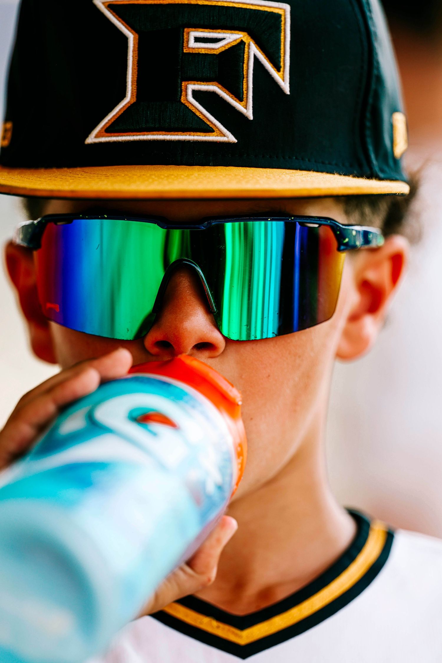 Baseball player wearing reflective sunglasses and team cap drinking from blue sports bottle.