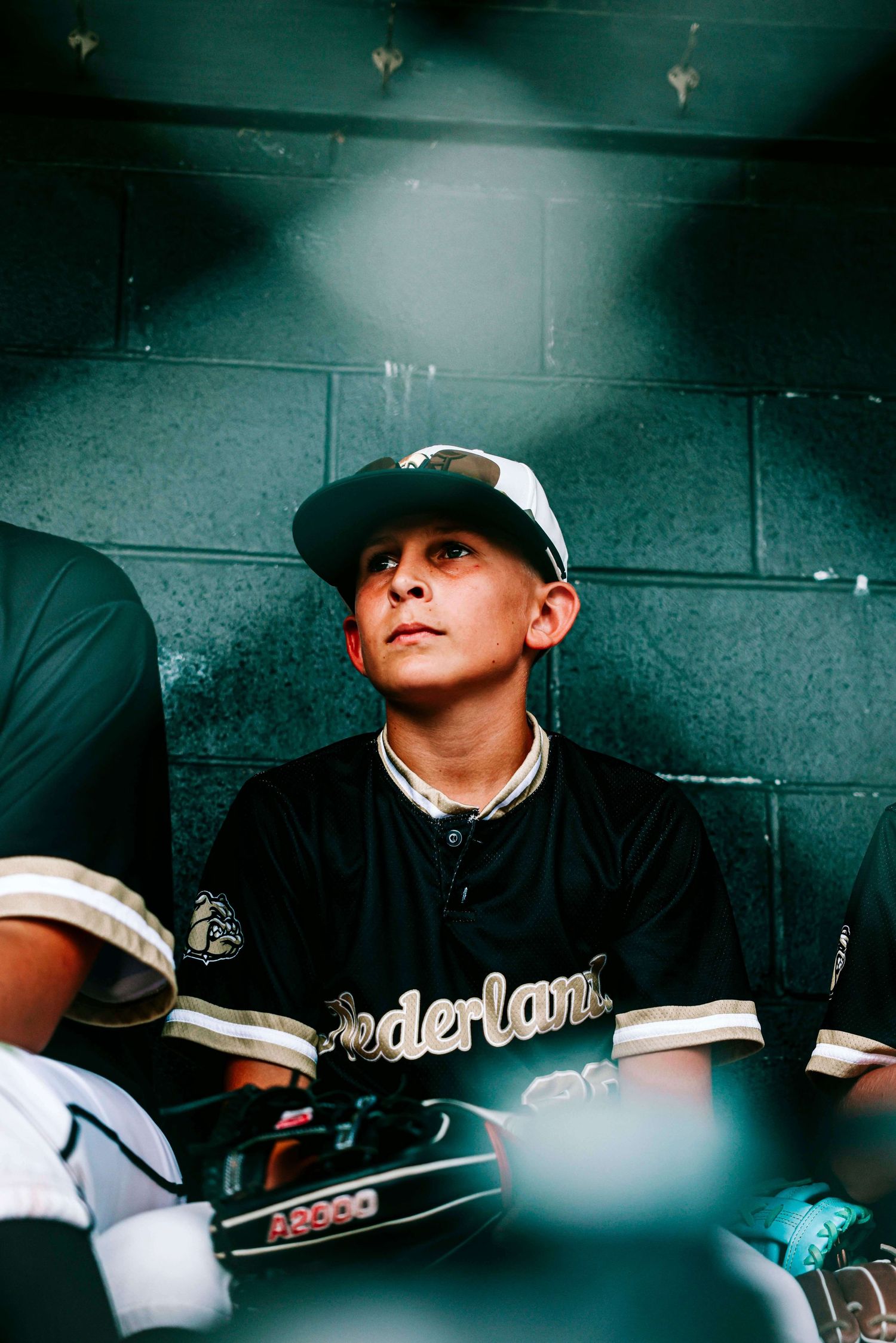 Baseball player in a dugout wearing a black uniform and white cap looks upward in dramatic lighting.