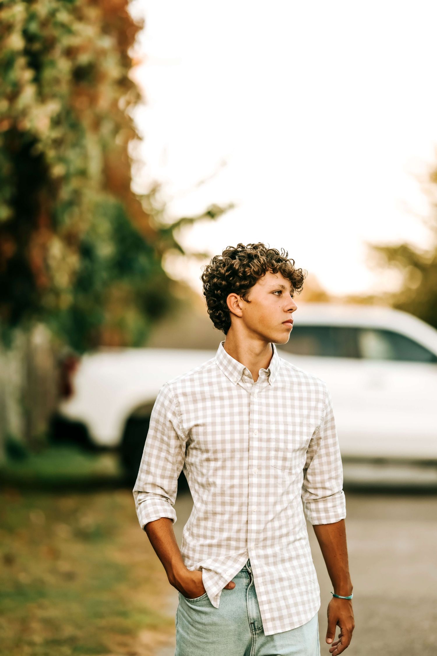 Person wearing checkered shirt poses casually with hands in pockets against outdoor backdrop.